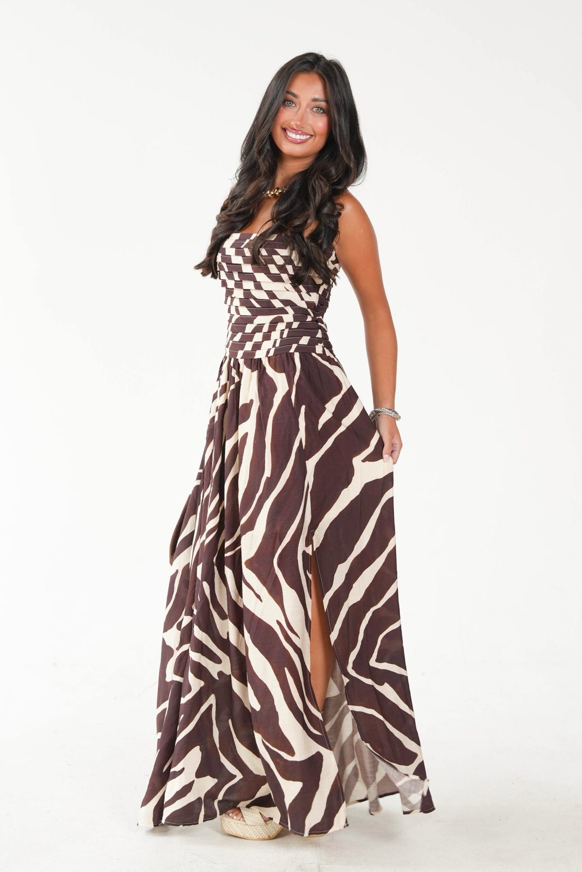 A woman modeling a sleeveless, floor-length maxi dress with a brown and beige zebra-like abstract pattern, standing against a plain white studio background.