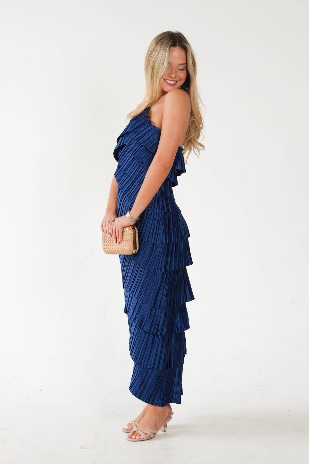 A woman modeling a navy blue pleated one-shoulder maxi dress with a ruffled tiered hem, holding a small beige clutch against a plain white studio background.