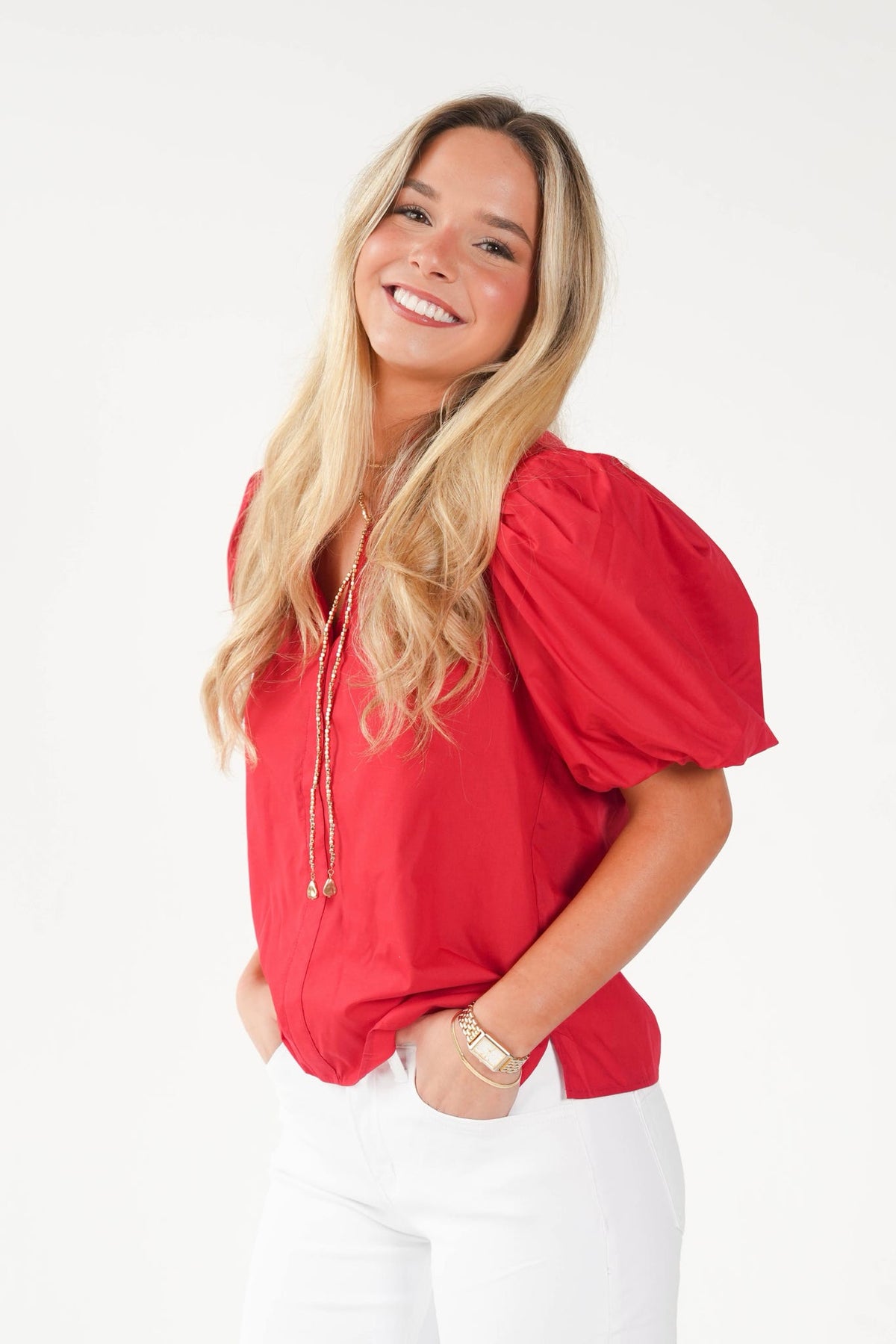 A smiling blonde woman wearing a bright red puff-sleeve blouse with a drawstring neckline and white pants, standing against a plain white studio background.