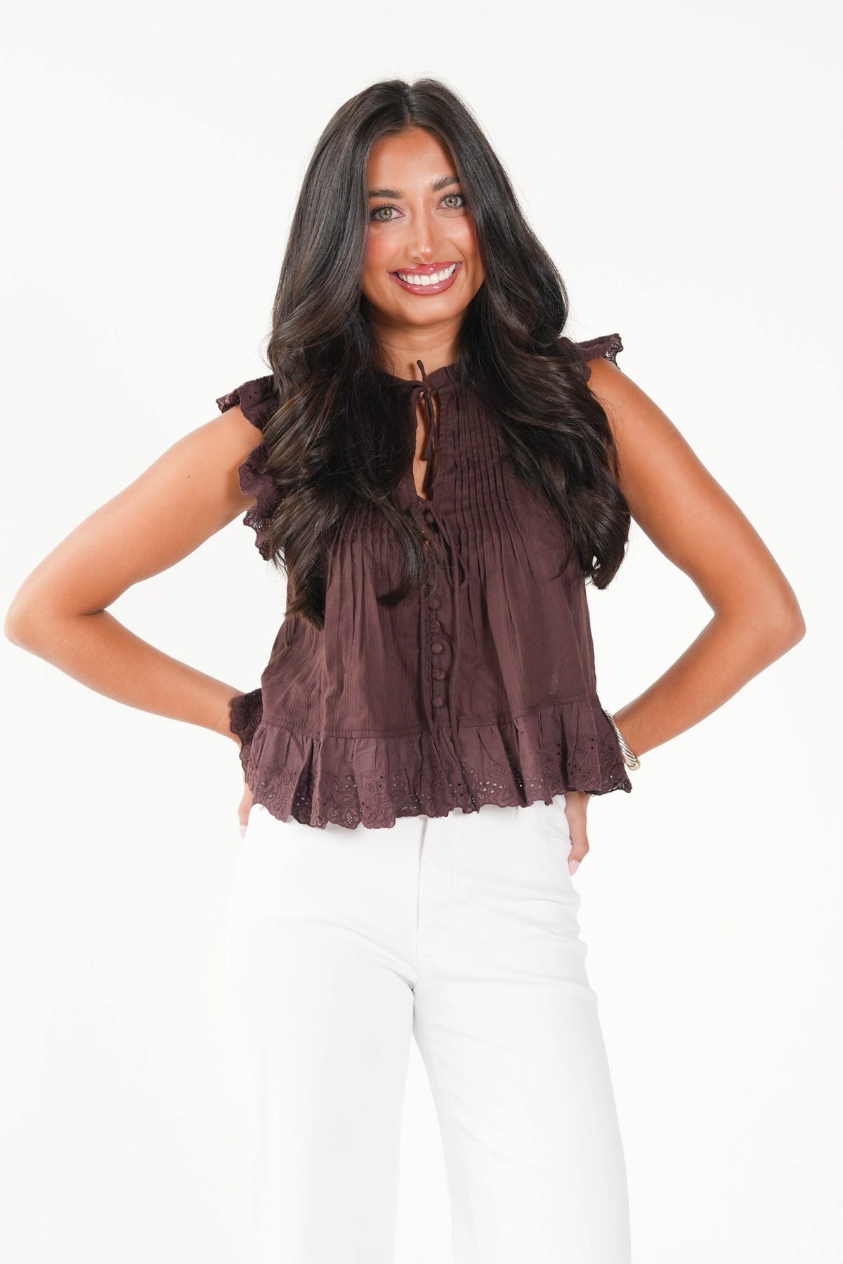 A woman modeling a sleeveless dark brown ruffled blouse with a tie-neck detail, paired with white pants, against a plain white background.