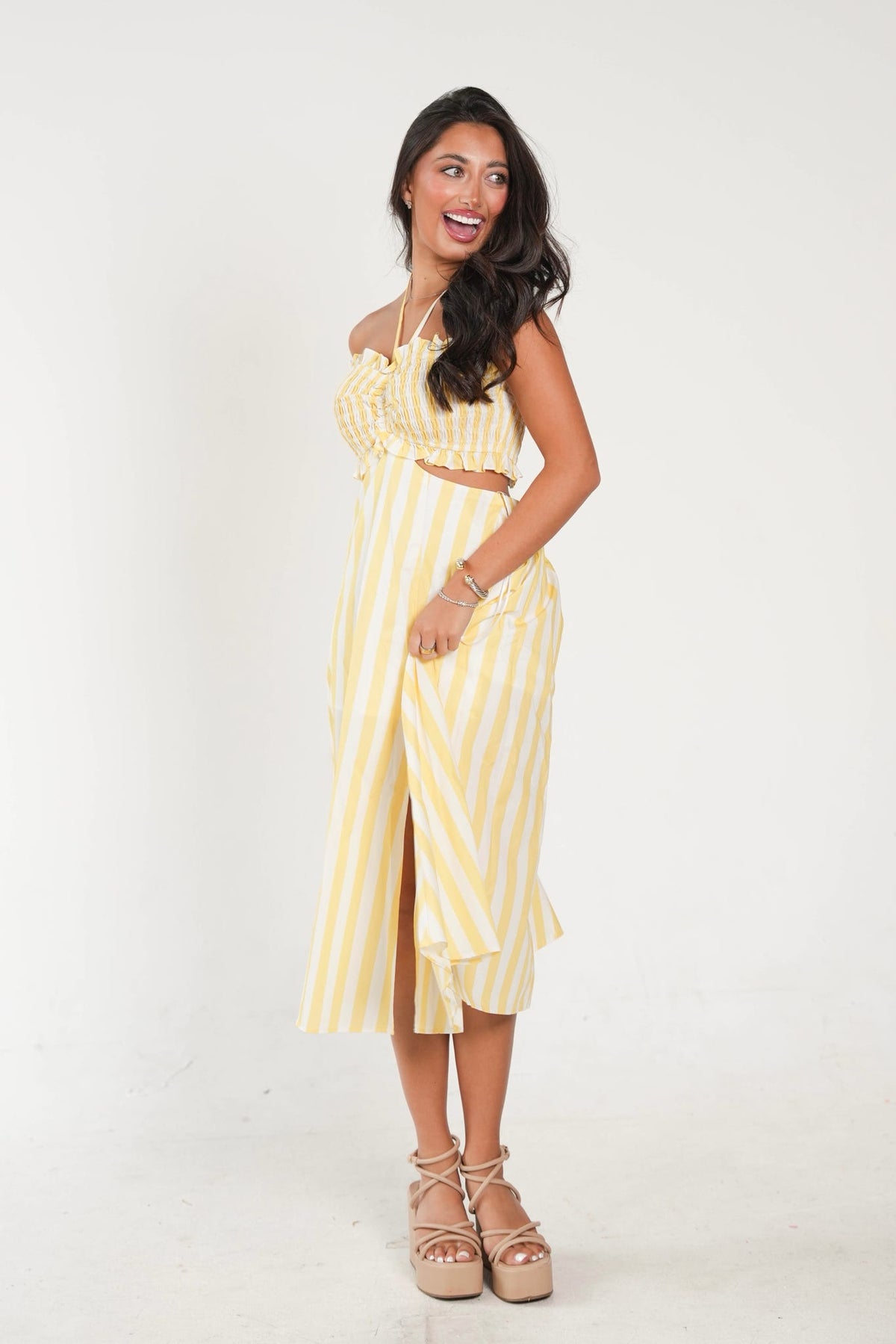 A woman modeling a yellow and white vertically striped midi dress with a halter top, side cutout, and front slit, paired with beige platform sandals against a plain white studio background.