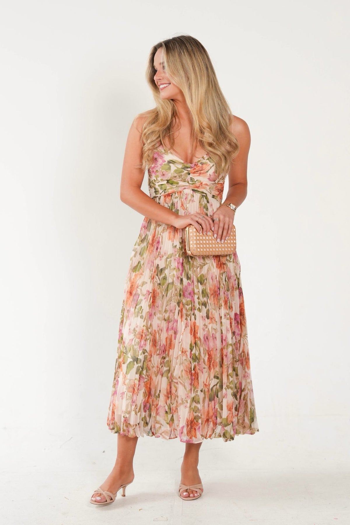 A woman modeling a sleeveless floral midi dress with a tie waist, holding a woven clutch and wearing clear high-heeled sandals against a plain white studio background.
