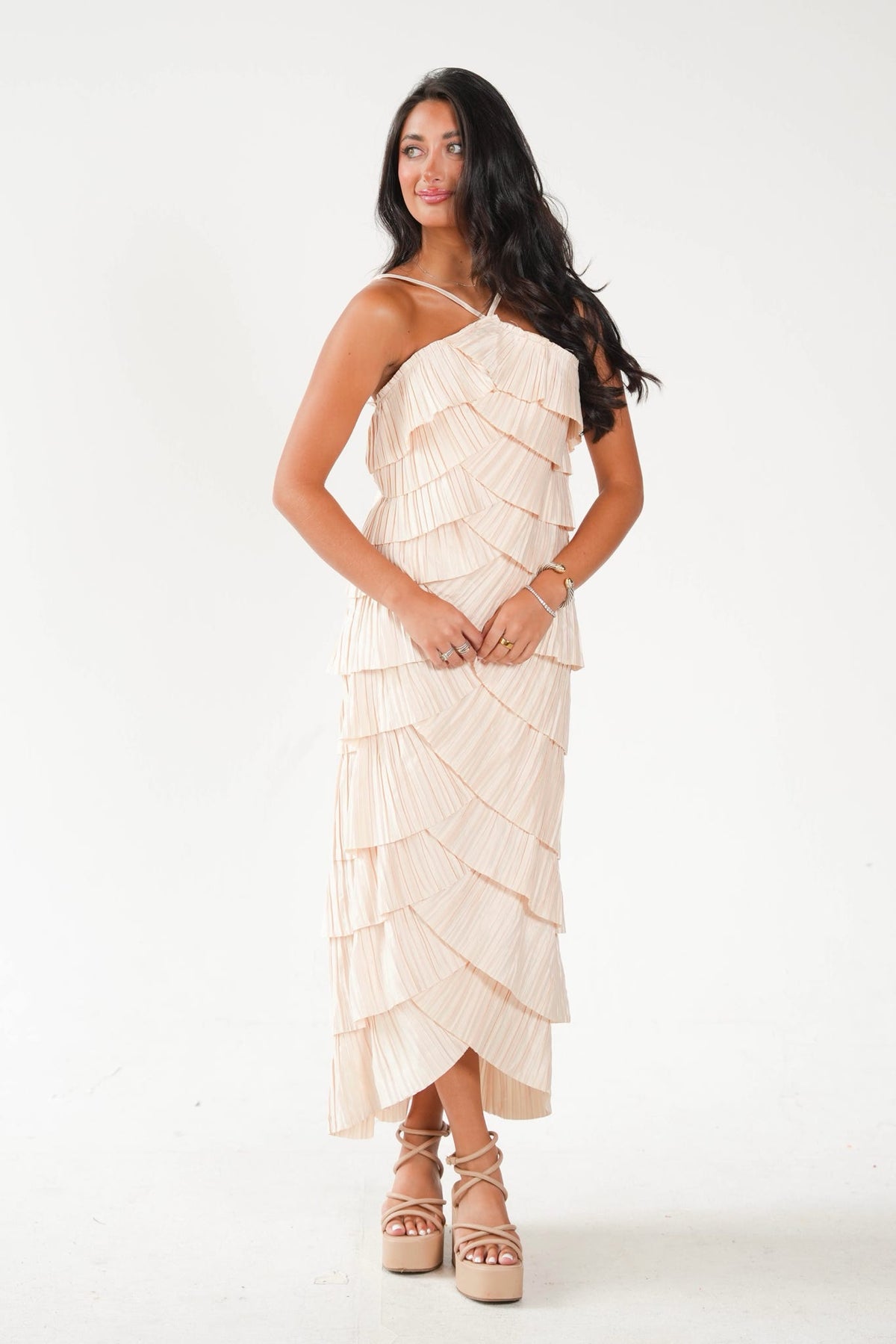 A woman modeling a one-shoulder, tiered maxi dress in a light cream or off-white color, paired with beige platform sandals, against a plain white studio background.