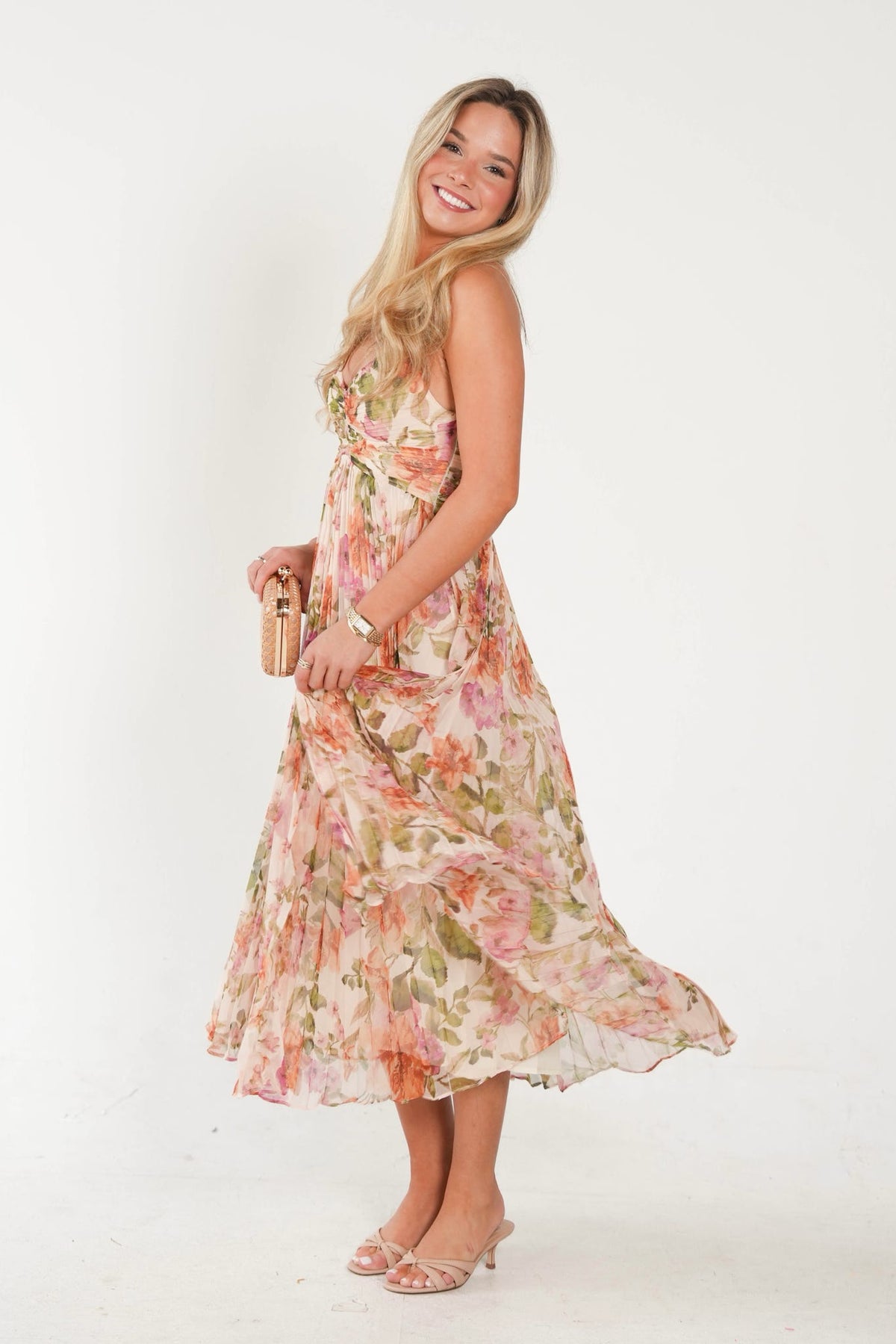 A woman modeling a sleeveless, tiered midi dress with a pink and green floral print on a cream background, holding a small gold clutch against a plain white studio backdrop.