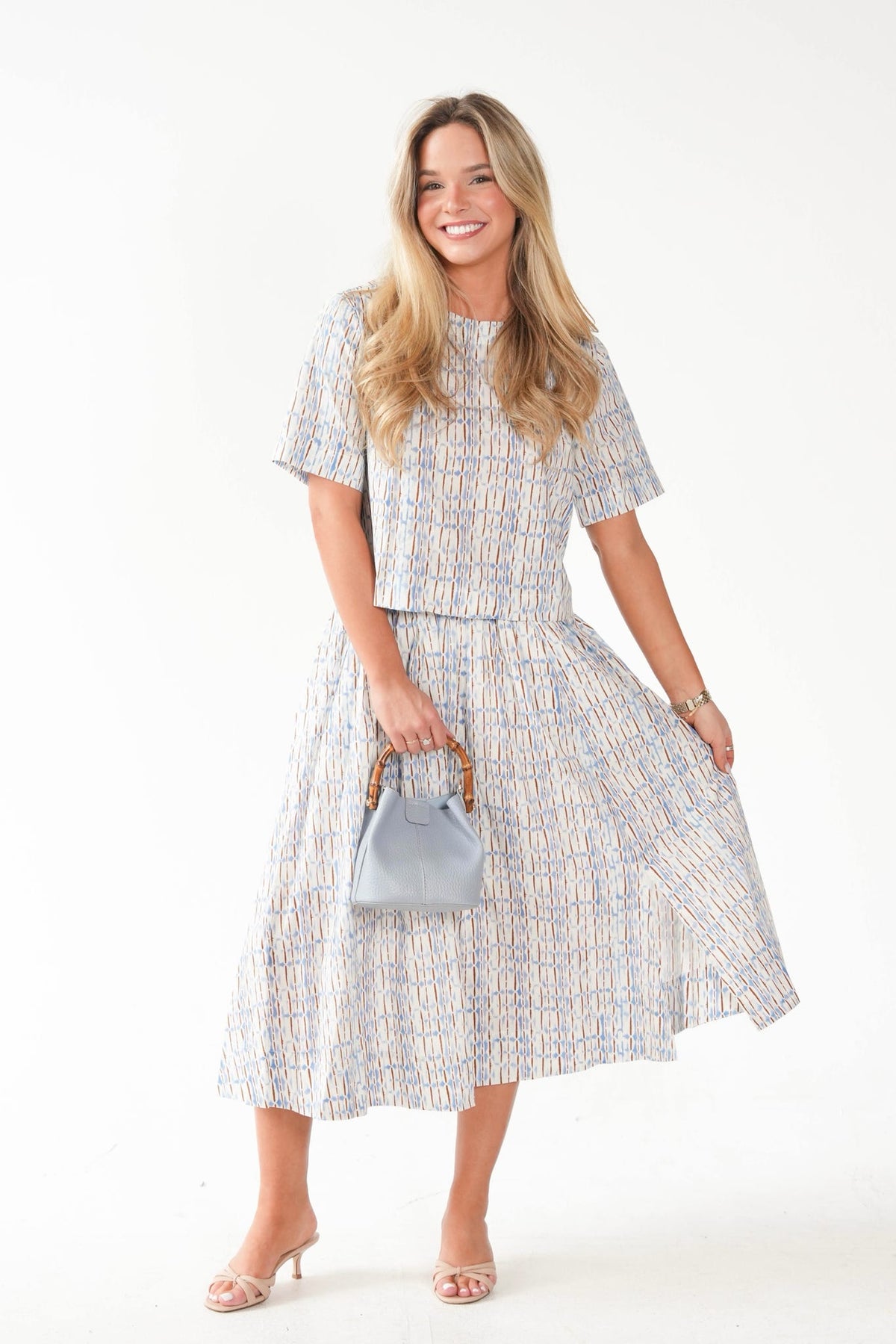 A woman modeling a matching short-sleeve cropped top and midi skirt set in a light blue, beige, and white geometric pattern, holding a small light blue handbag against a plain white studio background.