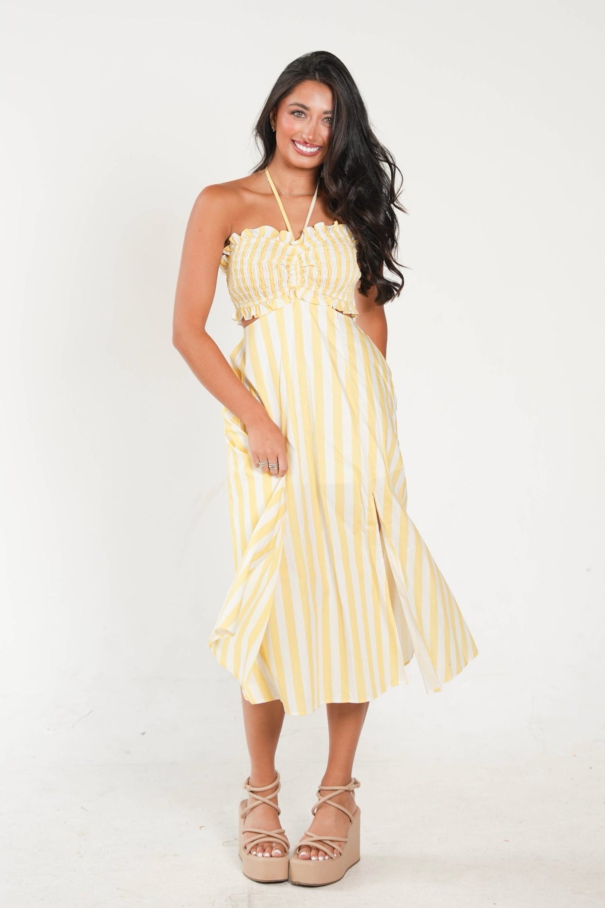 A woman modeling a yellow and white vertically striped strapless midi dress with a ruffled sweetheart neckline, gathered bodice, and draped skirt, paired with beige lace-up platform sandals against a plain white studio background.