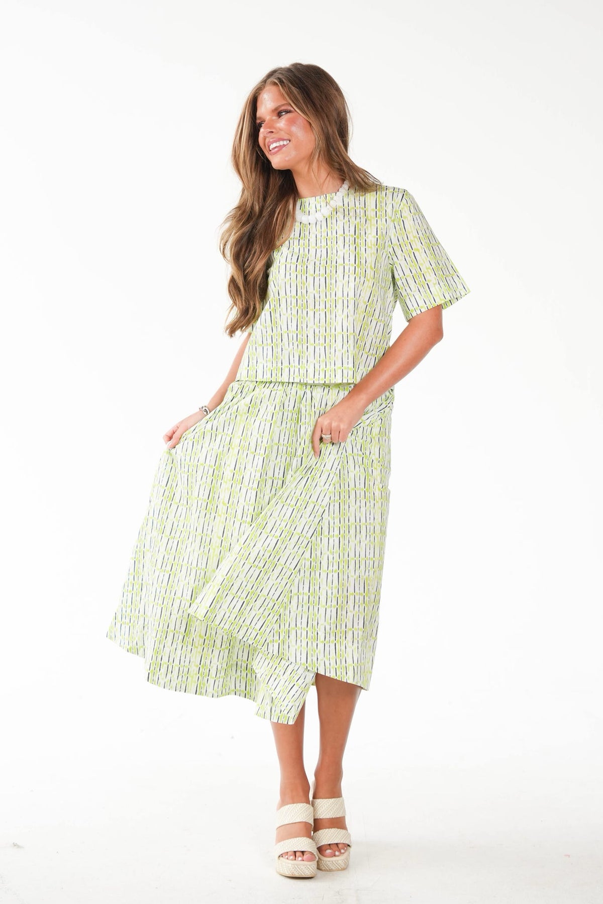 A woman modeling a light green and white vertically striped two-piece set consisting of a short-sleeve top and a matching midi skirt with pockets, paired with white platform sandals, against a plain white studio background.