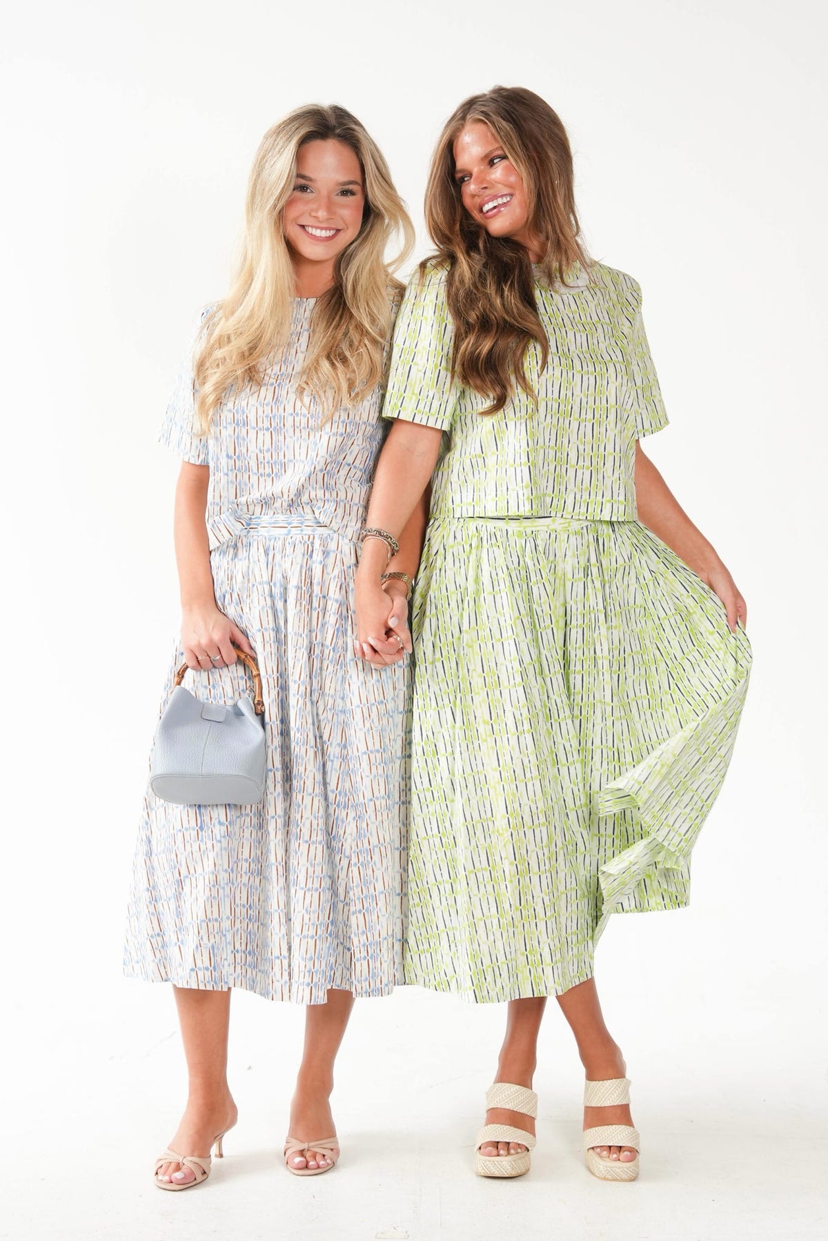 Two women modeling matching short-sleeve midi dresses with a vertical abstract stripe pattern. The woman on the left wears a light blue, white, and pink patterned dress and holds a small gray handbag.