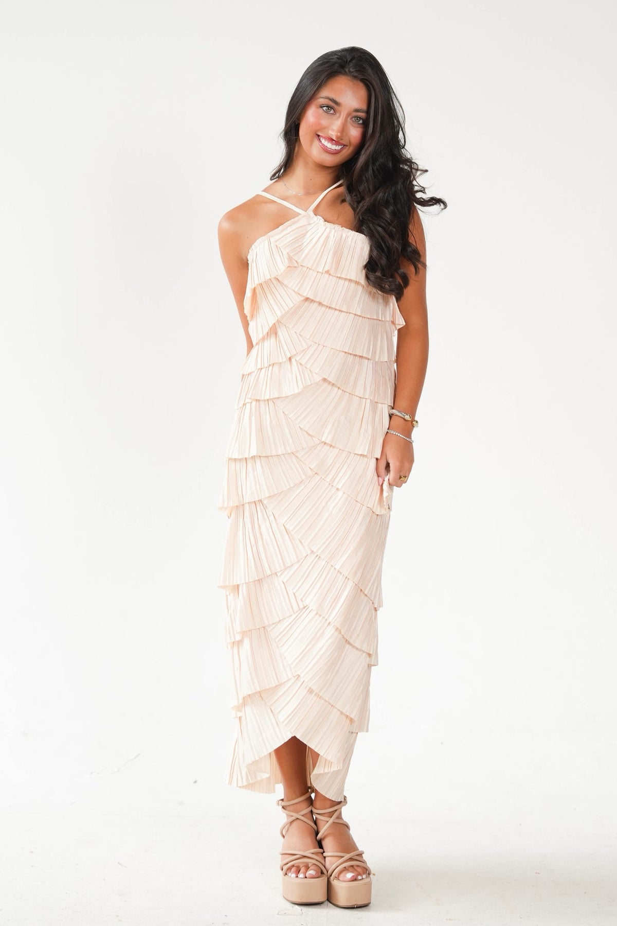 A woman modeling a light beige halter-neck midi dress with a tiered ruffle design, paired with beige platform sandals, against a plain white studio background.
