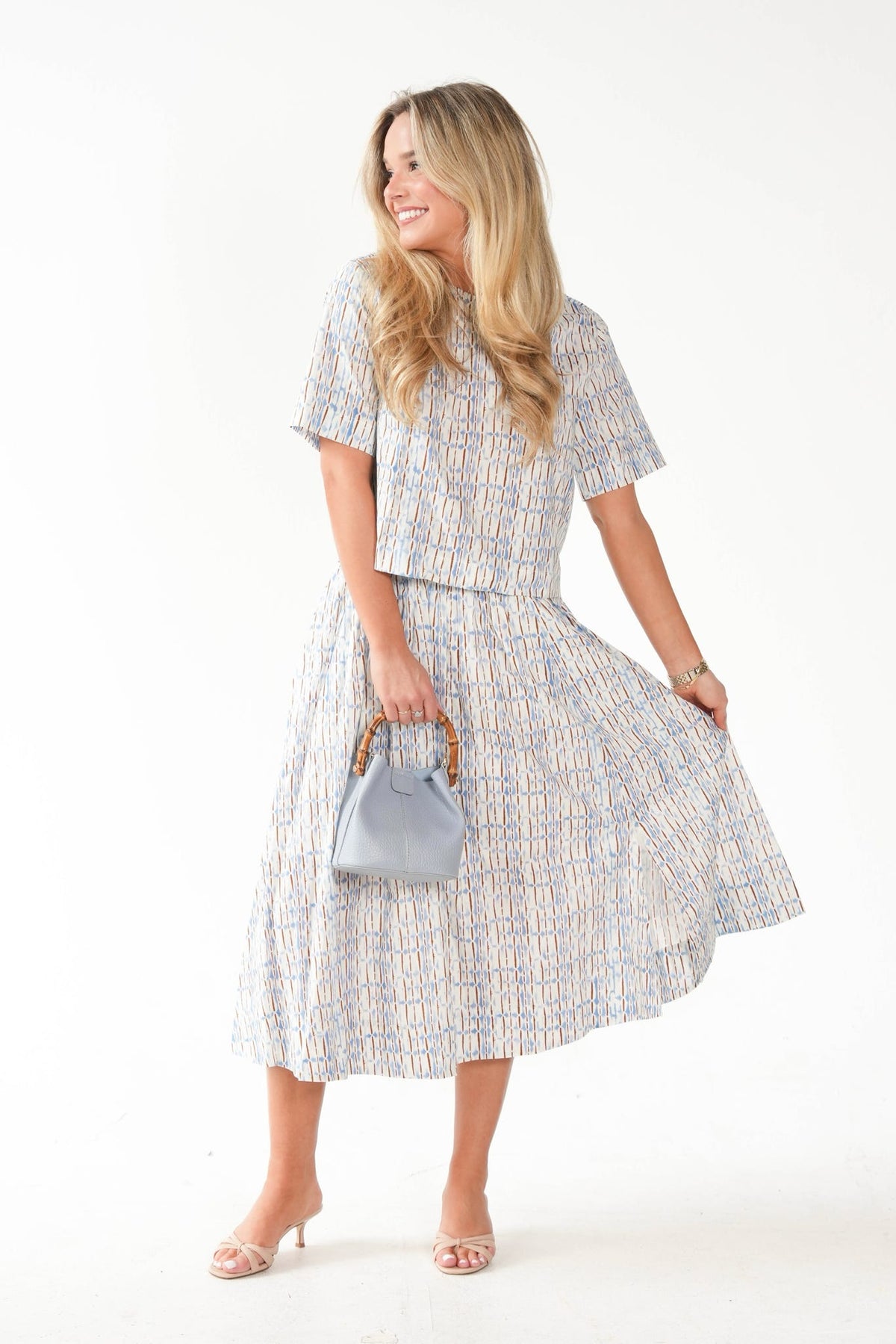 A woman modeling a matching short-sleeve top and midi skirt set in a light blue, white, and beige geometric pattern, holding a small light blue handbag against a plain white studio background.