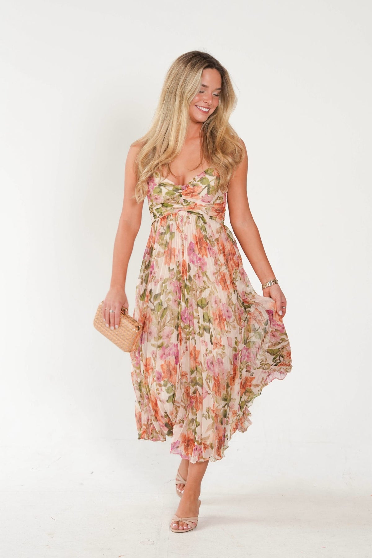 A woman modeling a sleeveless floral midi dress with a ruffled hem, holding a small woven clutch bag against a plain white studio background.