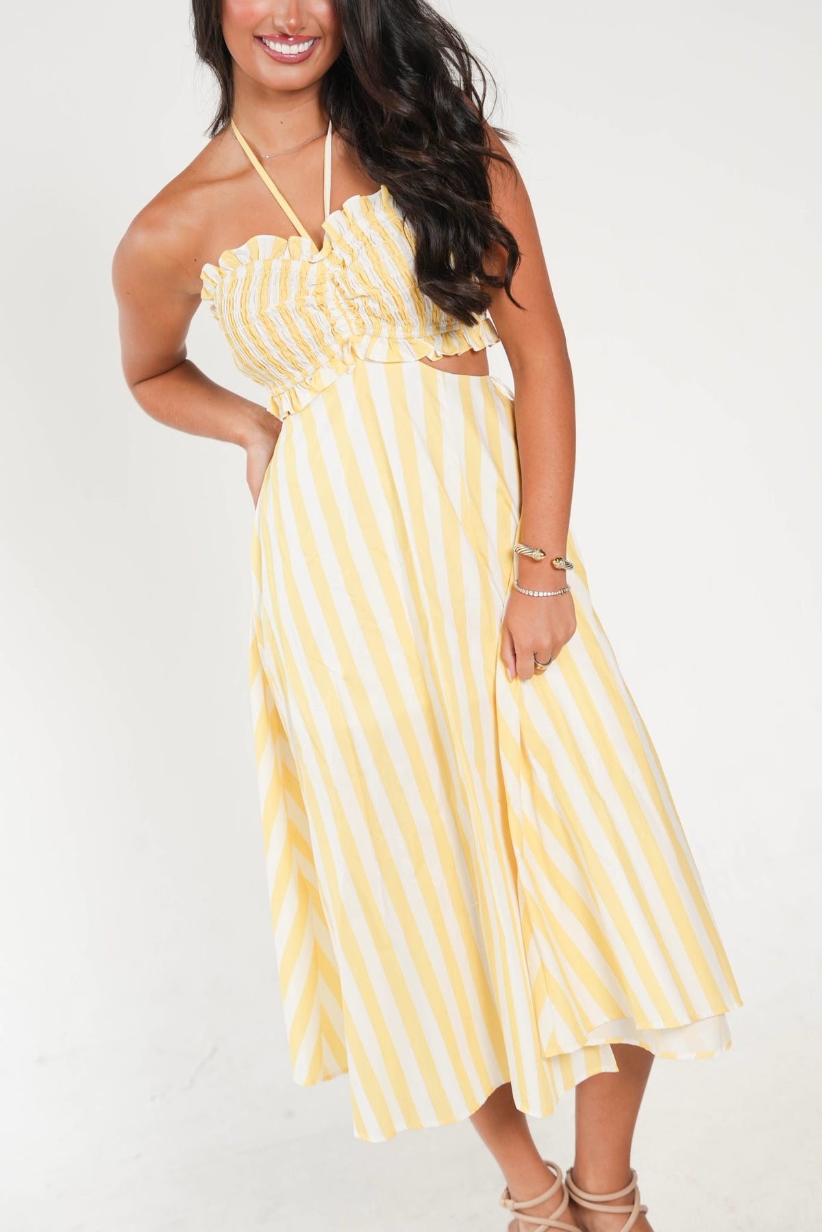 A woman wearing a yellow and white vertically striped halter midi dress with a ruched bust and side cutouts, paired with beige strappy high-heel sandals against a plain white studio background.