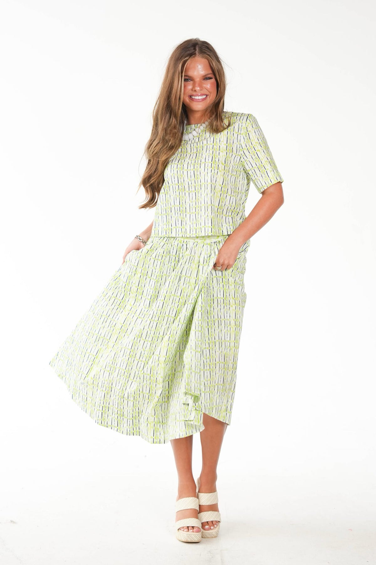 A woman modeling a light green and white vertically striped midi dress with short sleeves, a high neckline, and side pockets, paired with beige platform sandals against a white studio background.