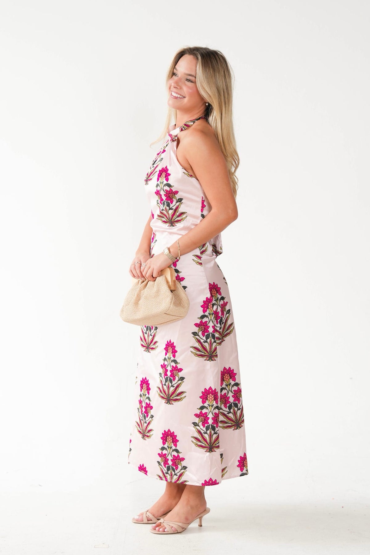 A woman modeling a sleeveless halter-neck maxi dress with a pink floral block print on a light pink base, holding a beige clutch bag against a plain white studio background.