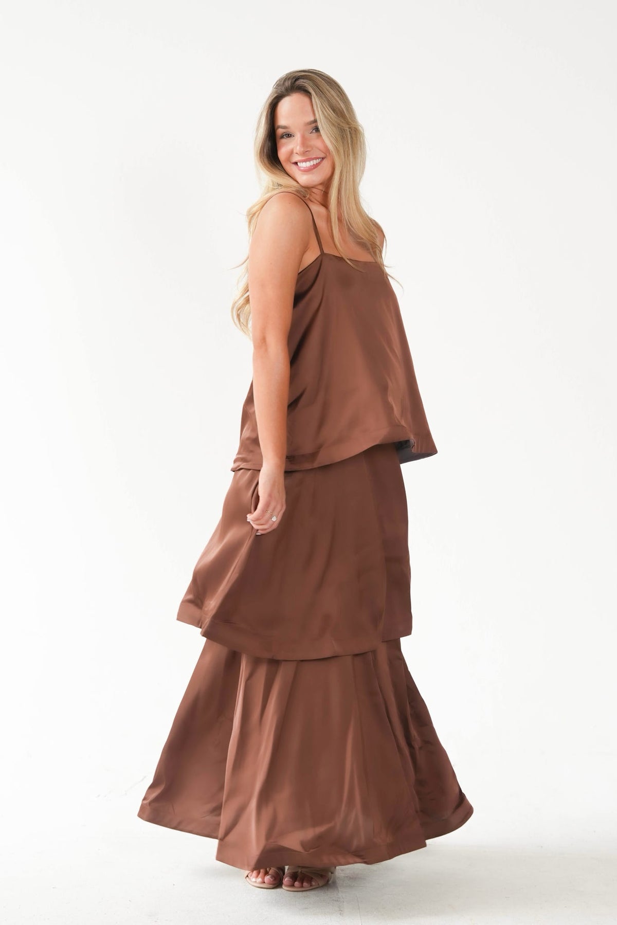 A woman modeling a matching two-piece lounge set in a warm brown color, featuring a sleeveless top and a tiered maxi skirt, against a plain white studio background.