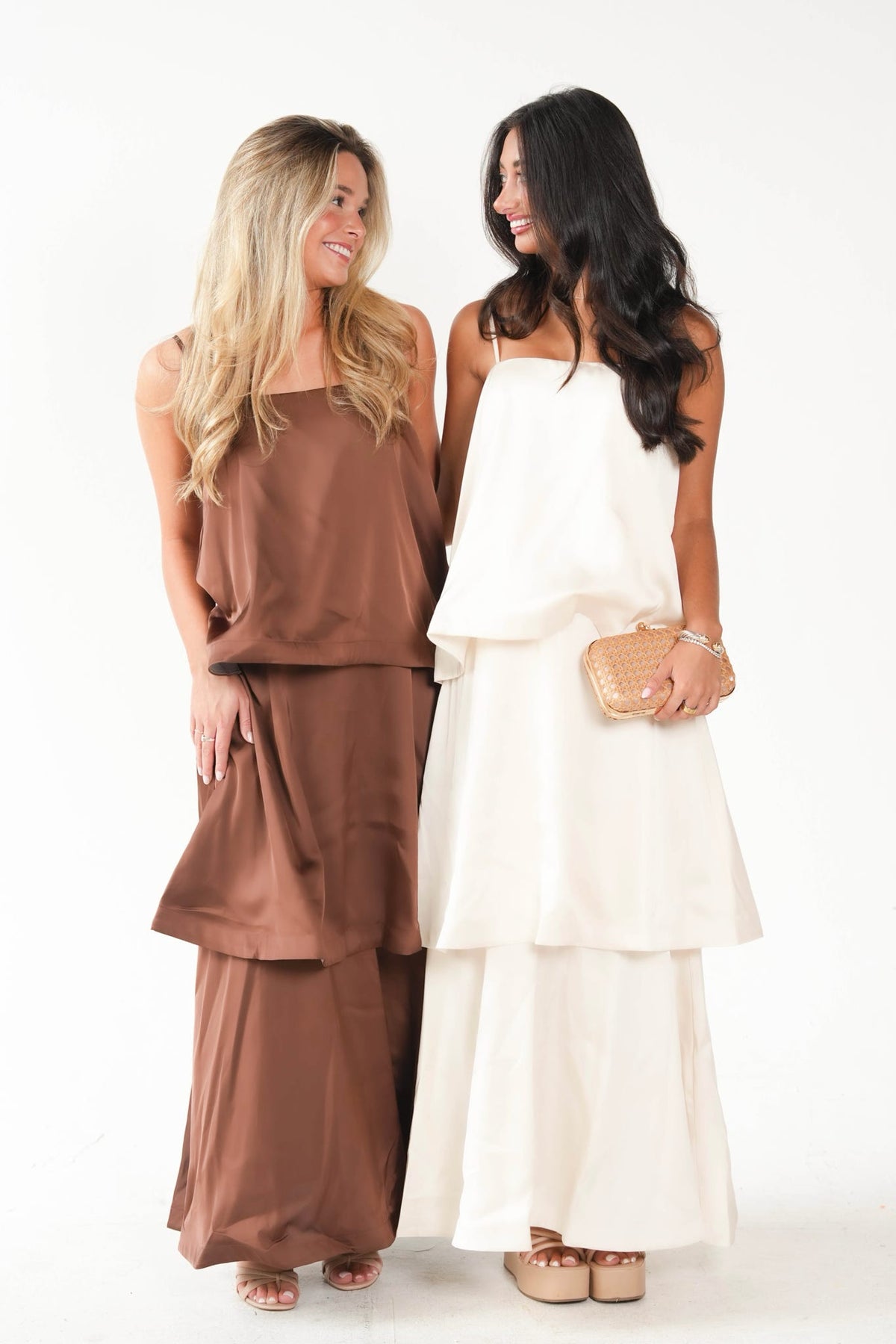 Two women standing side by side in a studio setting, wearing matching coordinated outfits. The woman on the left wears a brown sleeveless top and wide-leg pants set, and the woman on the right wears an off-white sleeveless tiered dress.