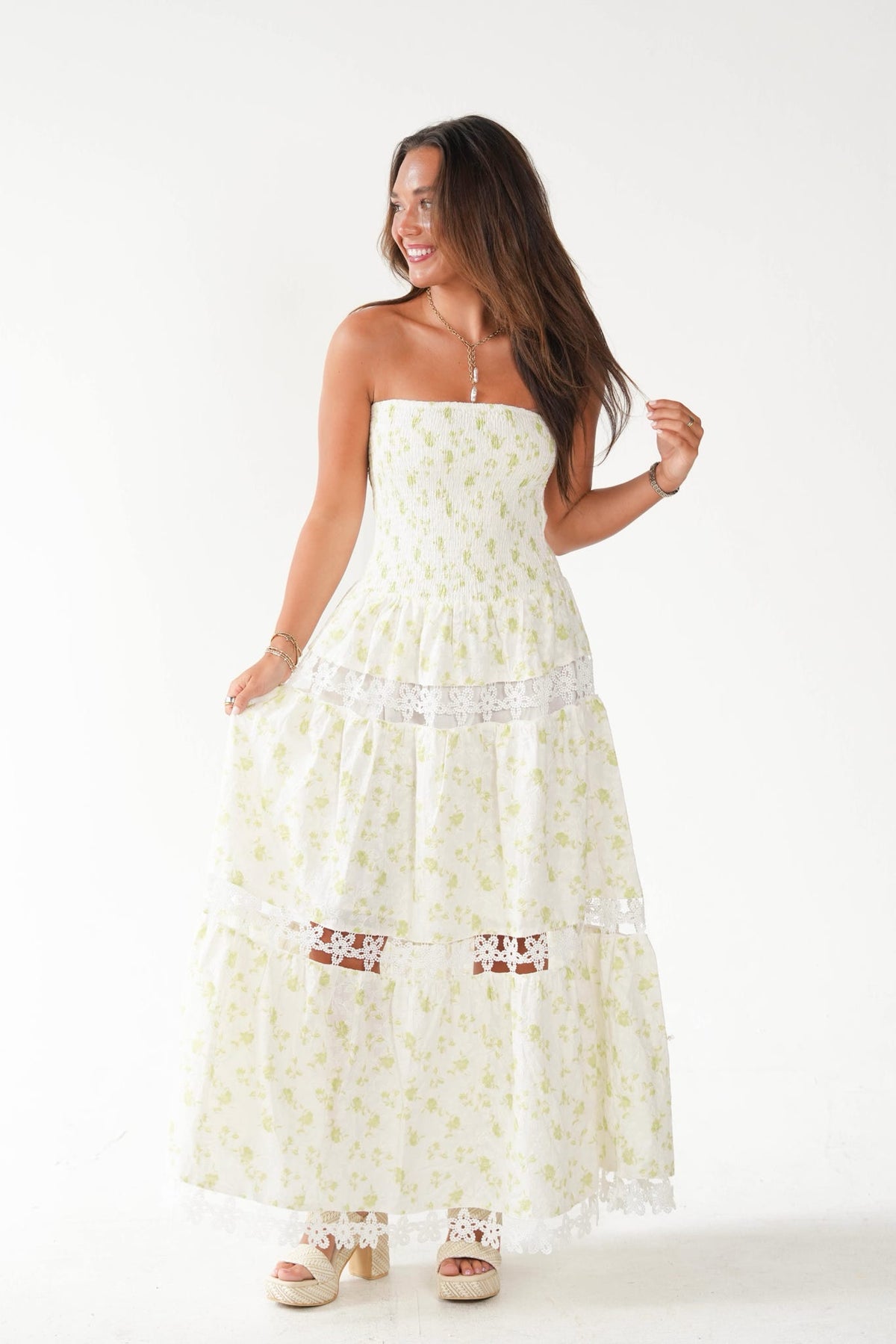 A woman modeling a strapless tiered maxi dress with a light yellow and white floral print, lace trim, and a white embroidered band at the waist, set against a plain white studio background.