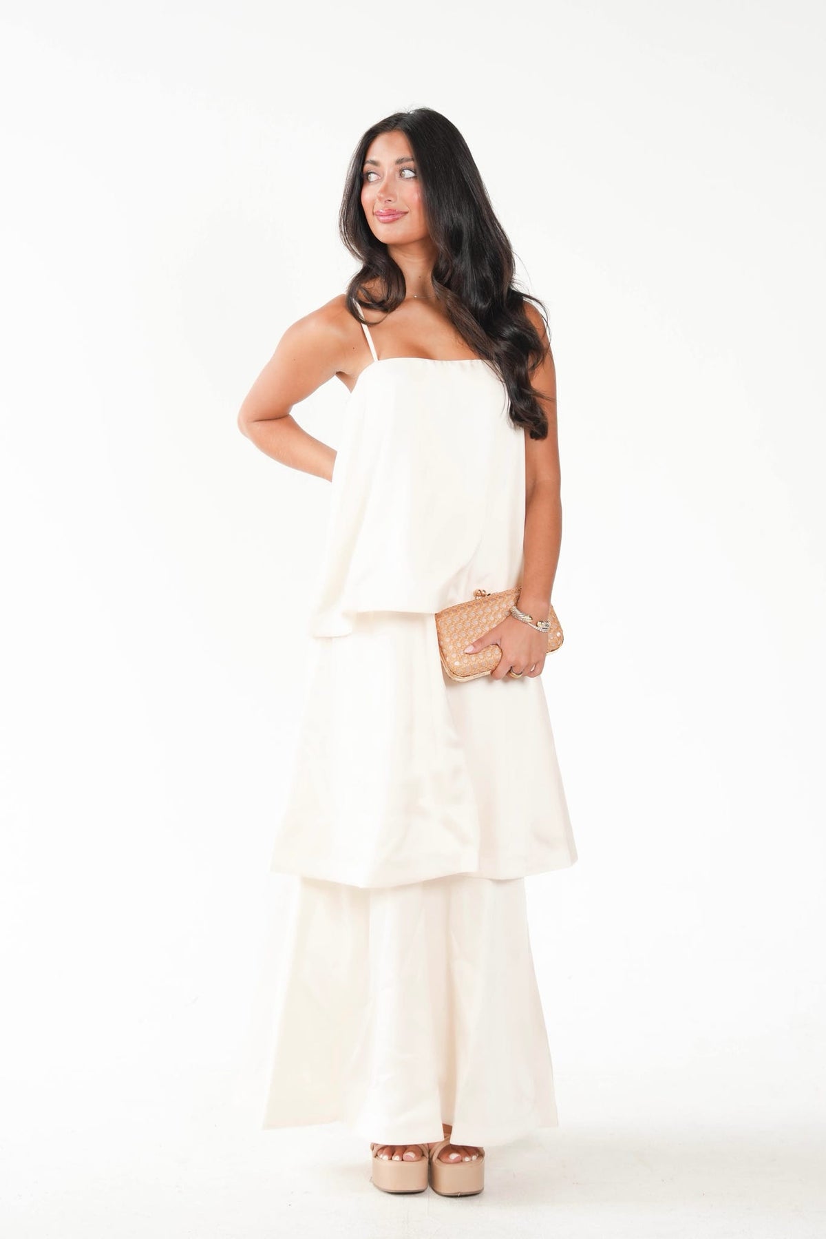 A woman modeling a white two-piece outfit consisting of a sleeveless tiered top and matching wide-leg pants, holding a small woven clutch against a plain white studio background.