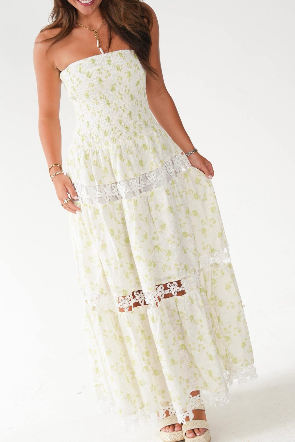 A woman wearing a strapless maxi dress with a light green floral print on a white background, featuring lace trim details, paired with beige platform sandals.