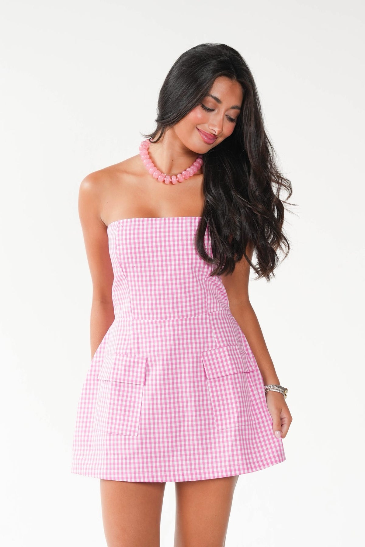 A woman modeling a pink and white gingham strapless mini dress with a flared skirt and front pockets, paired with a pink beaded necklace, against a plain white background.