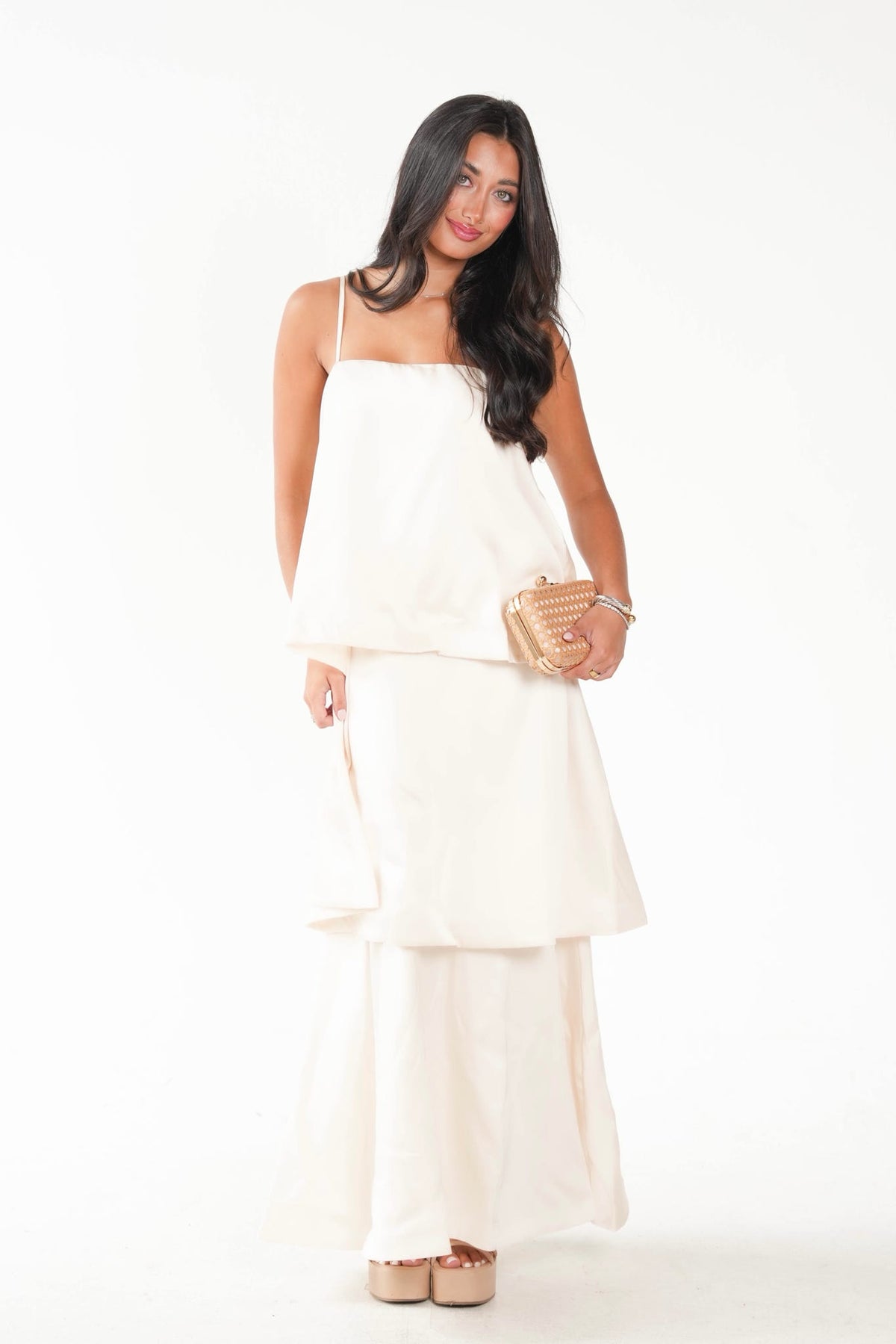 A woman modeling a cream-colored two-piece outfit consisting of a sleeveless tiered top and matching wide-leg pants, holding a small gold clutch against a plain white background.