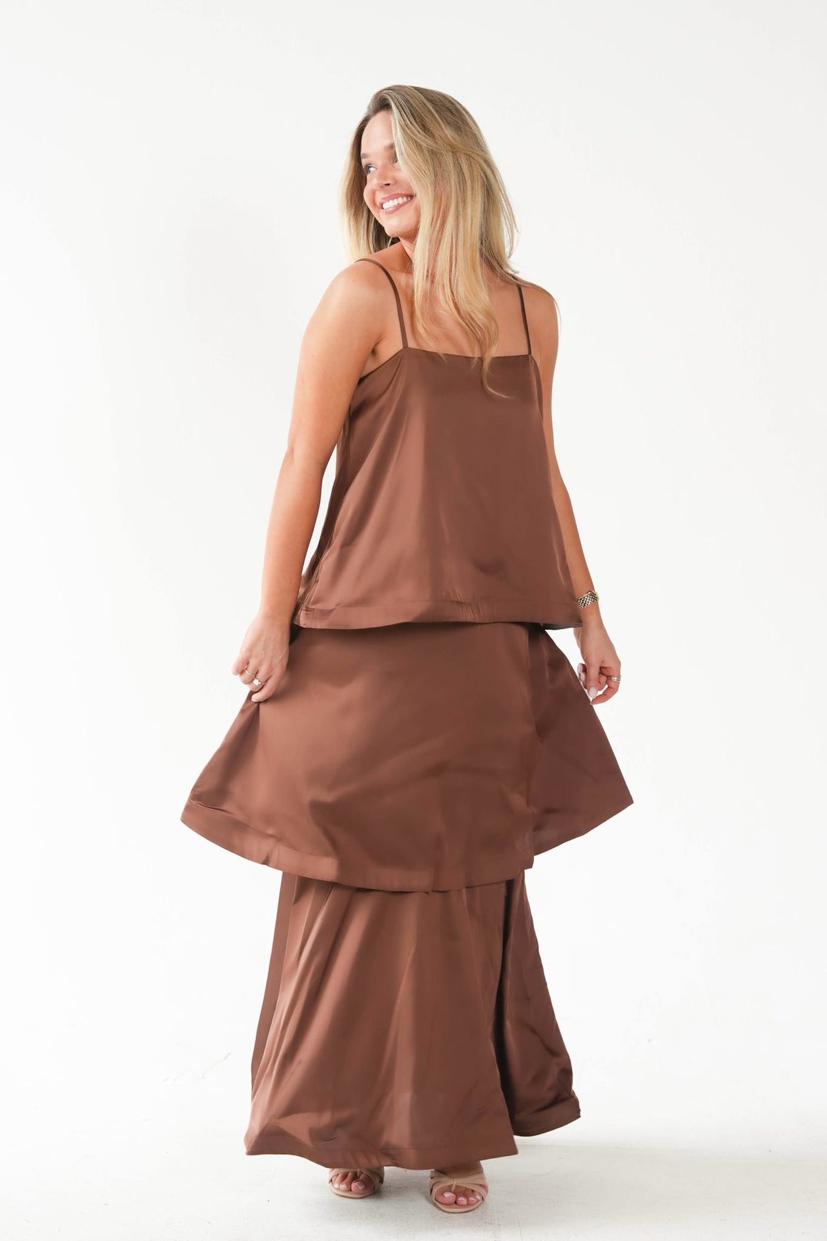 A woman modeling a two-piece brown satin-like outfit consisting of a spaghetti-strap camisole top and matching wide-leg pants, shown from a back-three-quarter angle against a plain white studio background.
