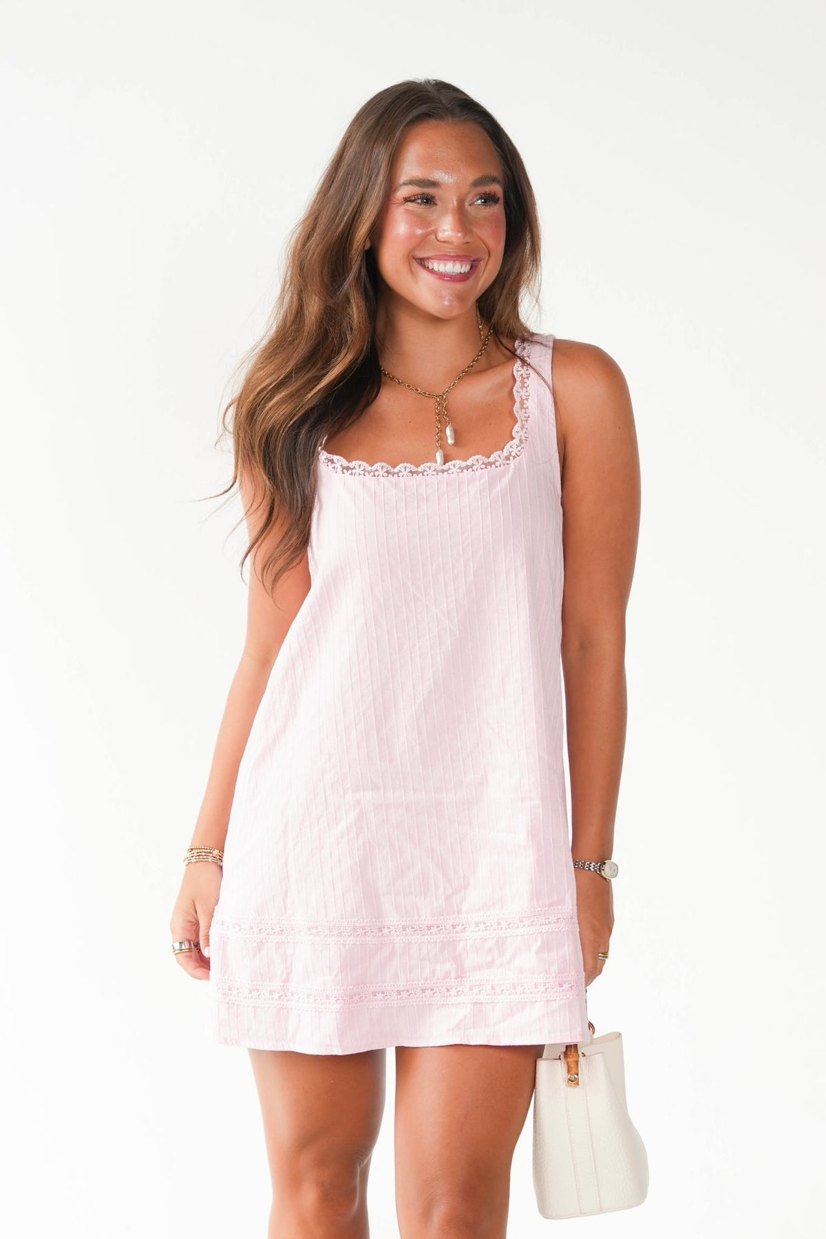 A woman modeling a light pink sleeveless cotton mini dress with scalloped lace trim on the neckline and hem, paired with a small white handbag against a plain white background.