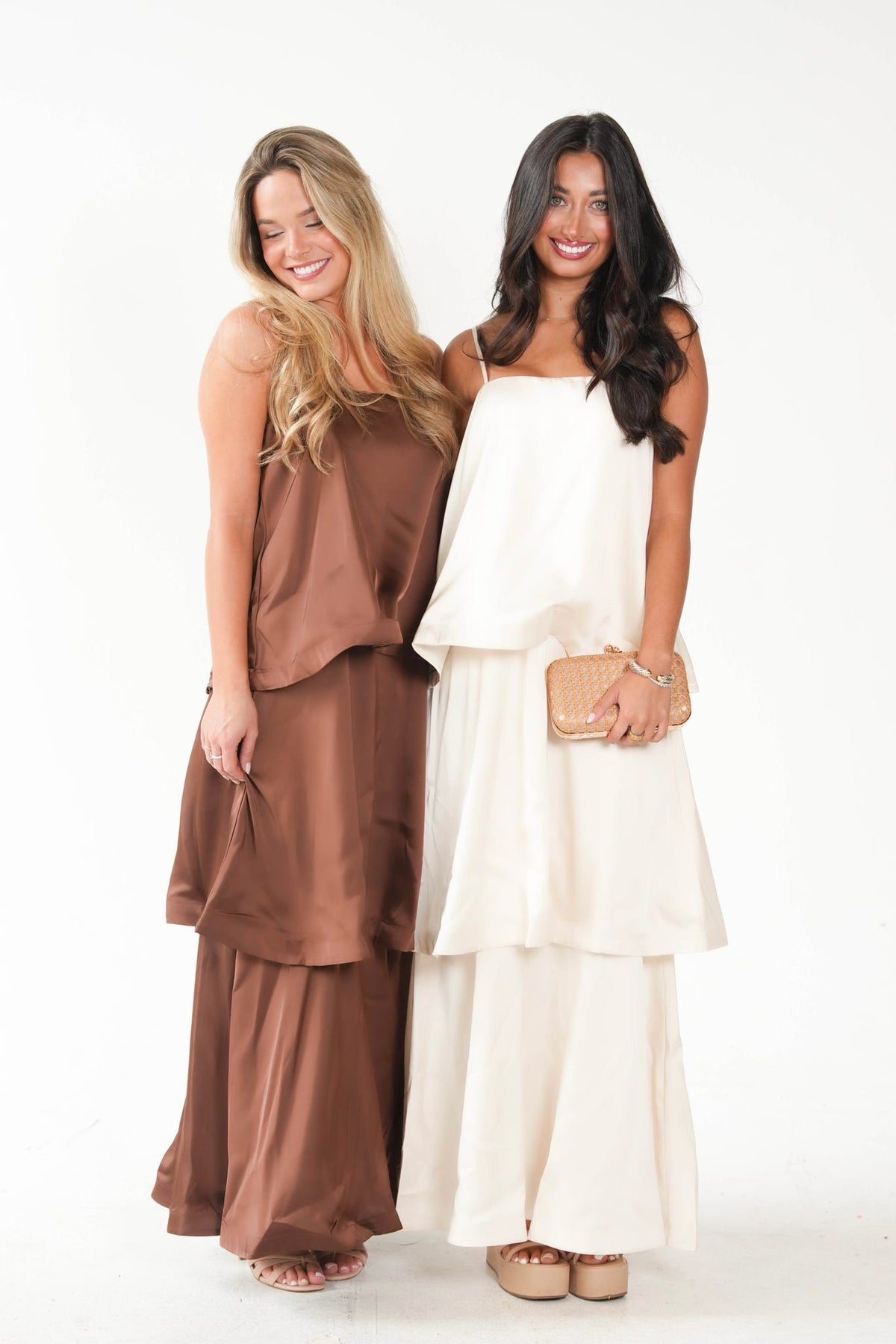 Two women modeling matching two-piece outfits against a plain white studio background. The woman on the left wears a brown satin-like sleeveless top with matching wide-leg pants.