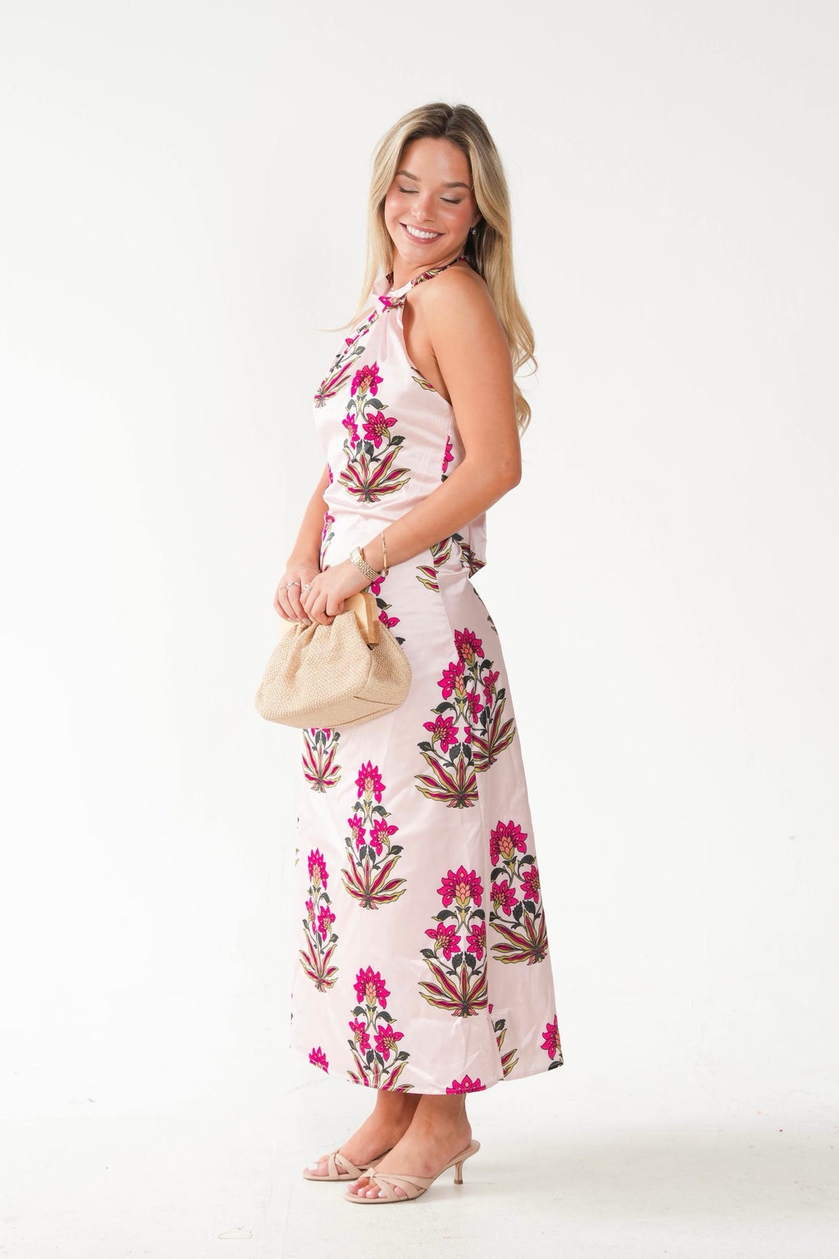 A woman modeling a sleeveless halter-neck maxi dress with a pink and green floral block print on a light pink base, holding a beige bucket bag against a plain white studio background.
