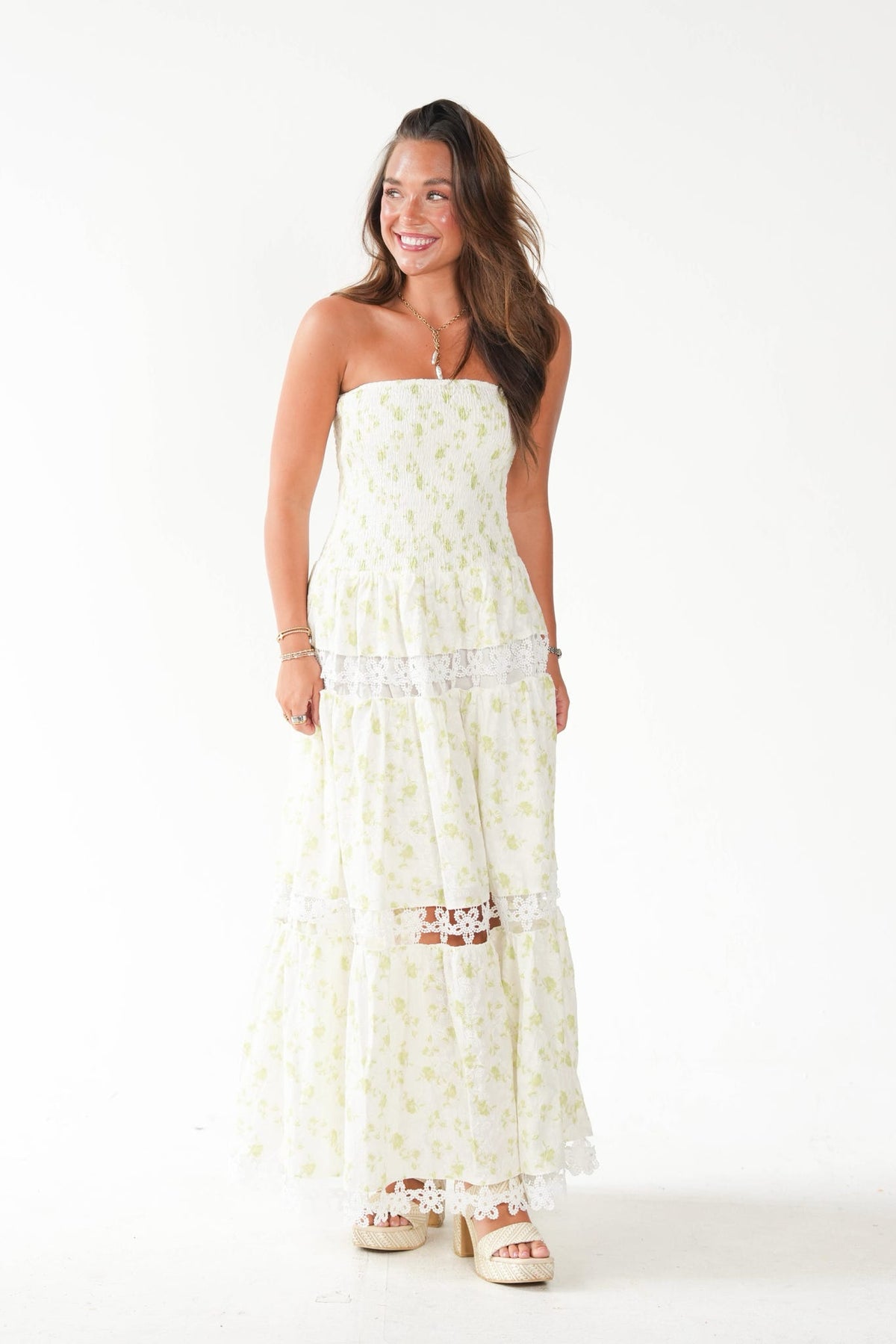A woman modeling a strapless maxi dress with a white base and light yellow floral print, featuring a lace-trimmed waistband and tiered skirt, set against a plain white studio background.