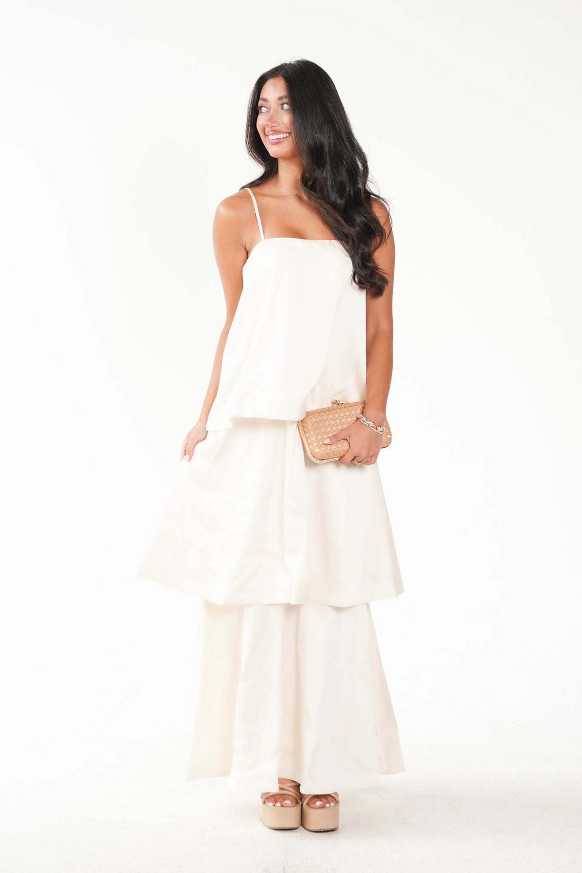 A woman modeling a two-piece cream or off-white satin set consisting of a spaghetti-strap top and matching wide-leg pants, styled with a woven clutch and platform sandals against a white studio background.