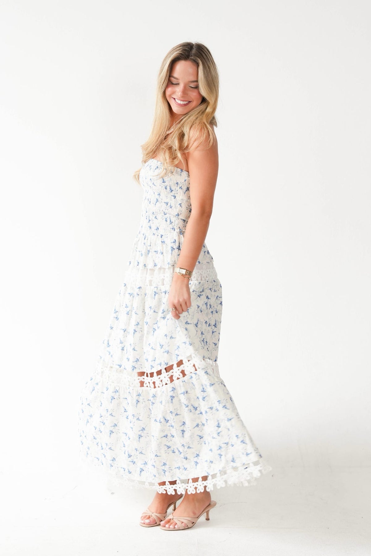 A woman modeling a white strapless maxi dress with a blue floral print and tiered ruffle hem, standing against a plain white studio background.