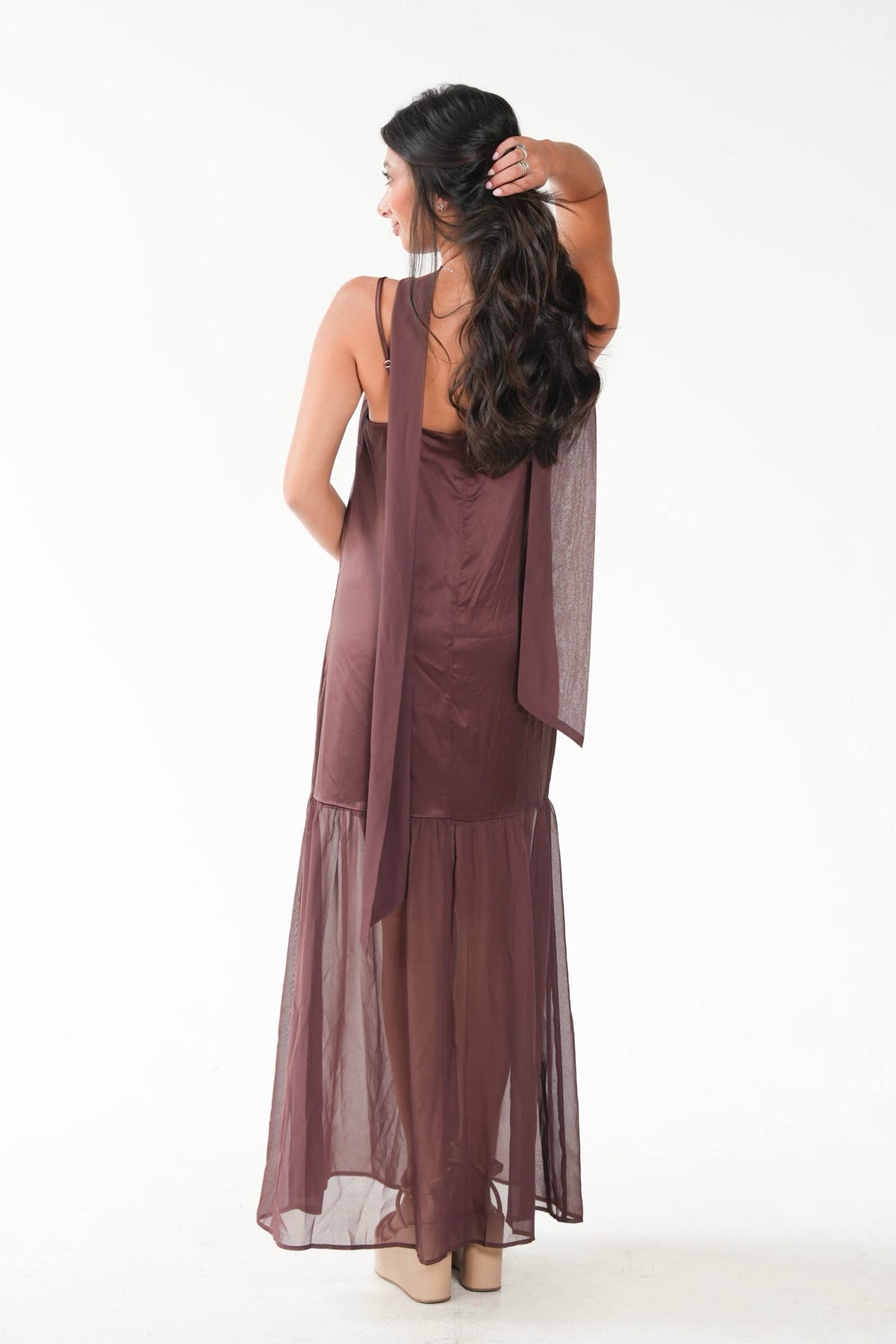 A woman viewed from the back wearing a sleeveless, floor-length mauve-brown dress with a sheer draped overlay and ruffled hem, photographed against a plain white studio background.
