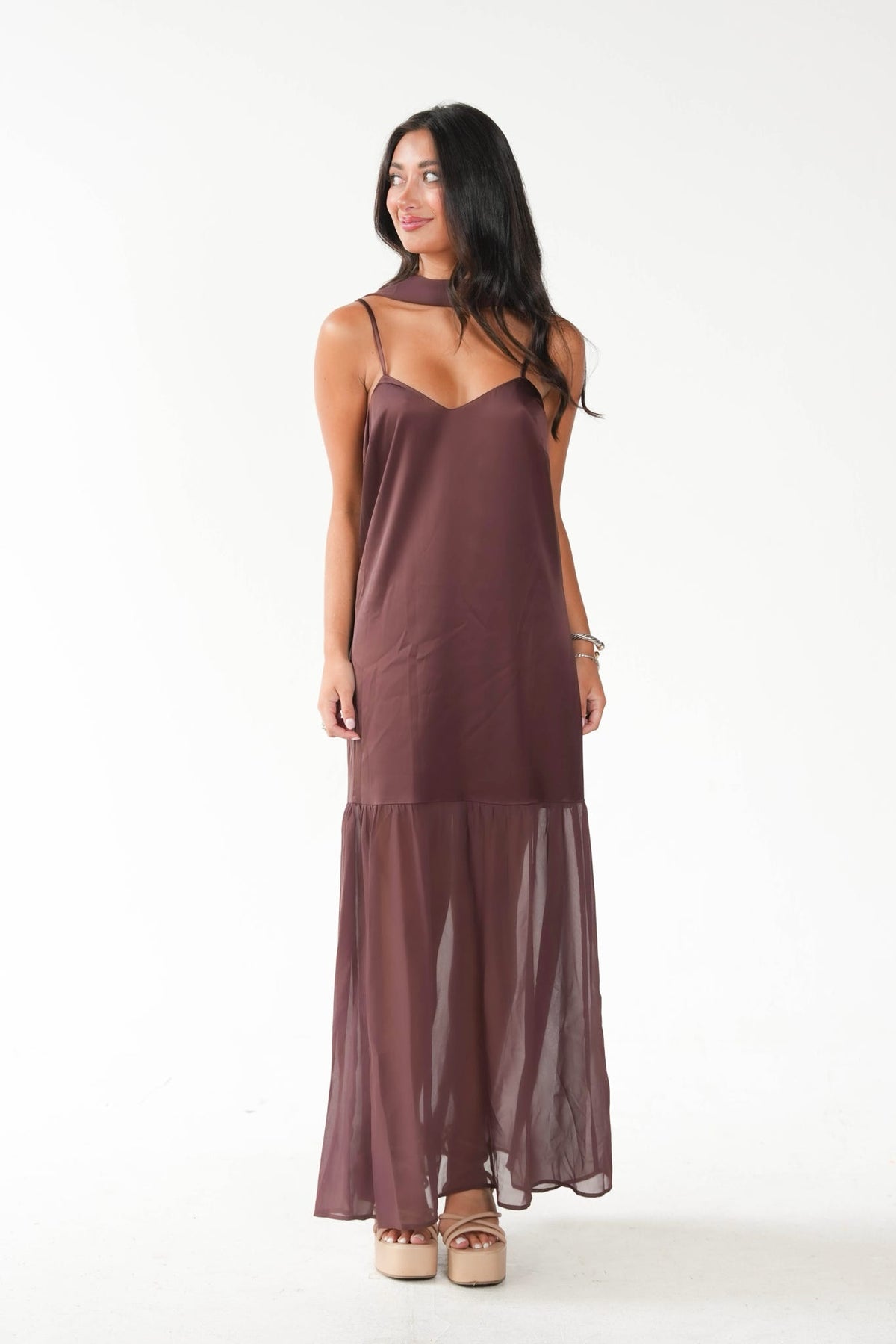 A woman modeling a sleeveless, high-neck maxi dress in a muted plum-brown color, featuring a sheer mesh tiered skirt, set against a plain white studio background.