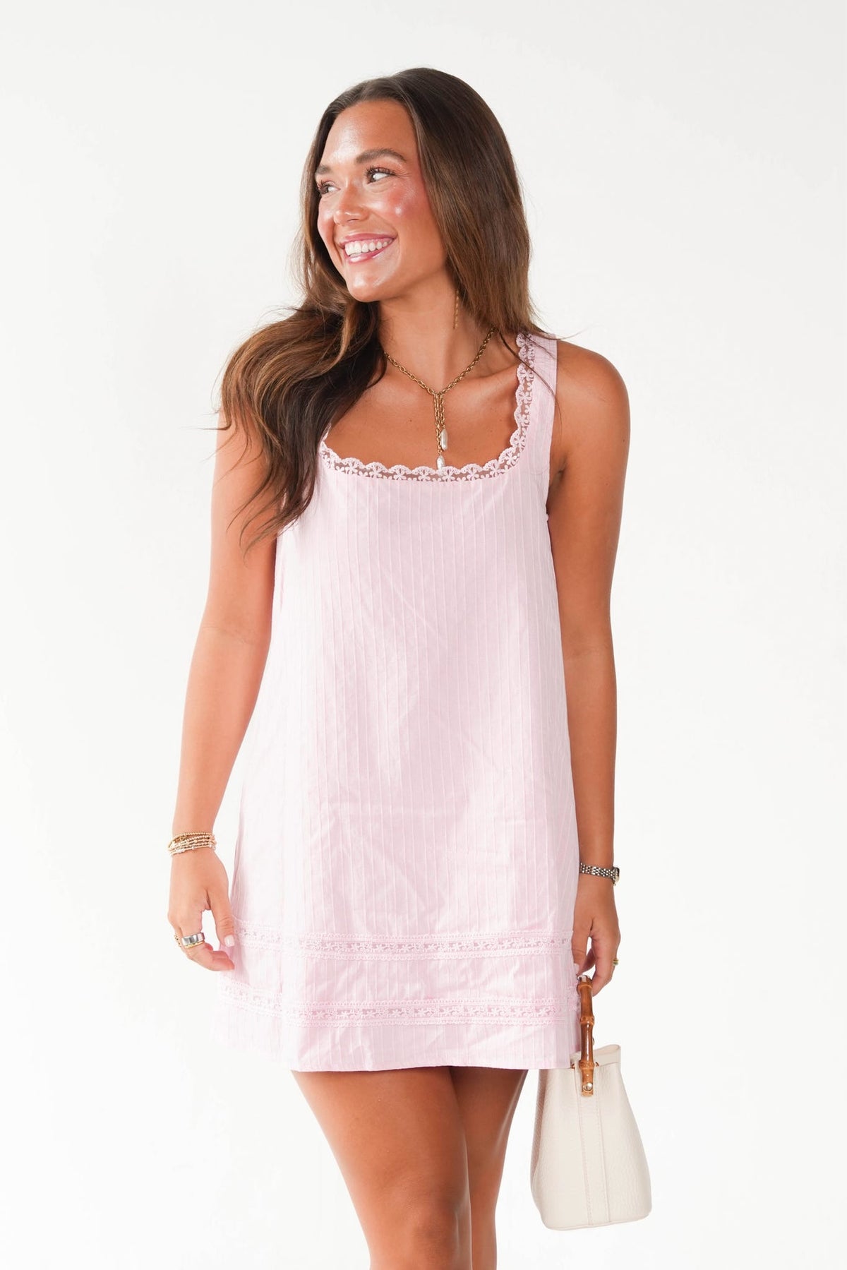A woman modeling a light pink sleeveless mini dress with delicate lace trim at the scoop neck and hem, holding a small white handbag against a plain white studio background.