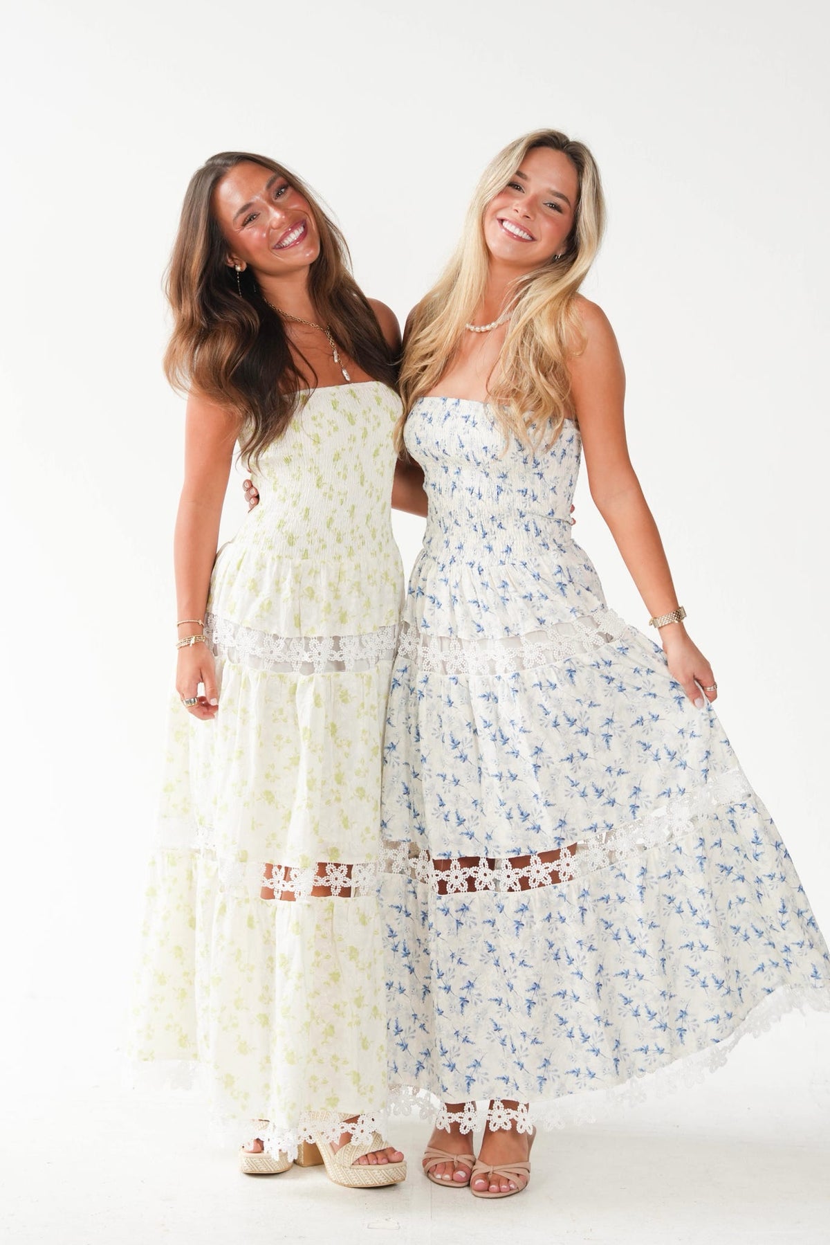 Two women standing side by side in strapless maxi dresses against a plain white studio background. The woman on the left wears a cream dress with a small yellow floral print, and the woman on the right wears a white dress with a small blue floral print.