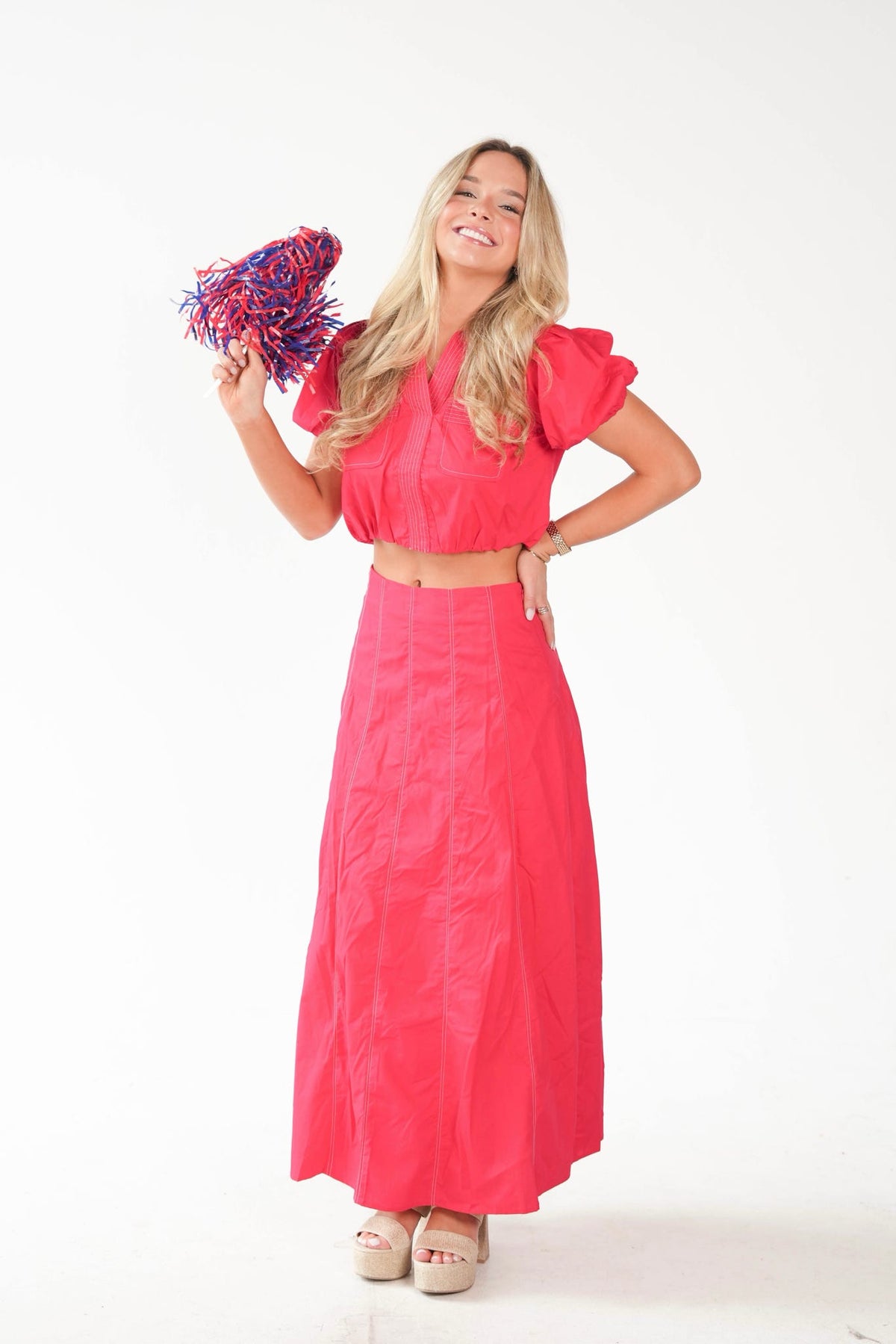 A woman modeling a bright pink two-piece outfit consisting of a short puff-sleeve crop top and a matching high-waisted maxi skirt, holding a blue and red pom-pom against a plain white background.