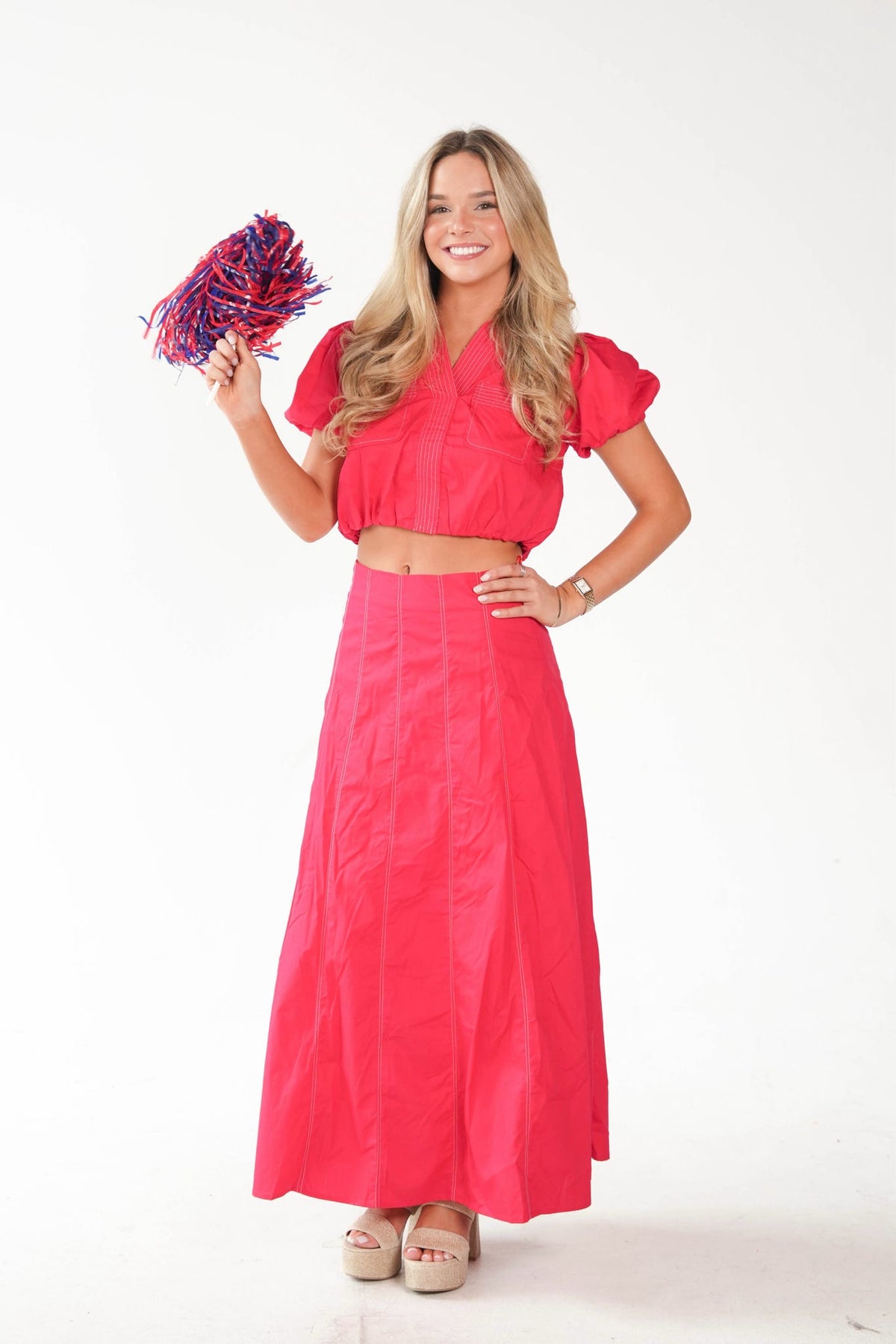 A woman modeling a bright pink two-piece outfit consisting of a short-sleeve crop top and a matching long skirt, holding a red and blue pom-pom against a plain white background.