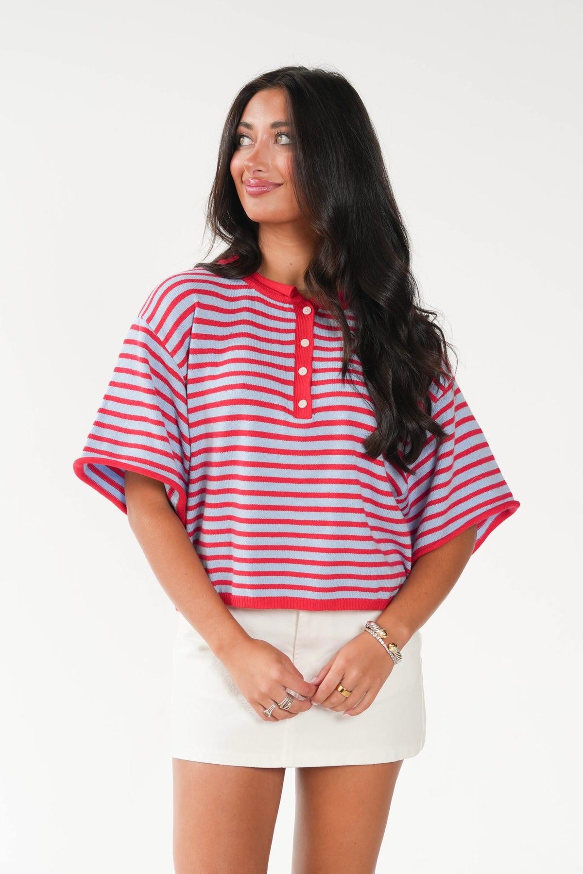 A woman modeling a short-sleeve striped henley top with red and white horizontal stripes and a red collar with three white buttons, paired with white shorts against a plain white background.