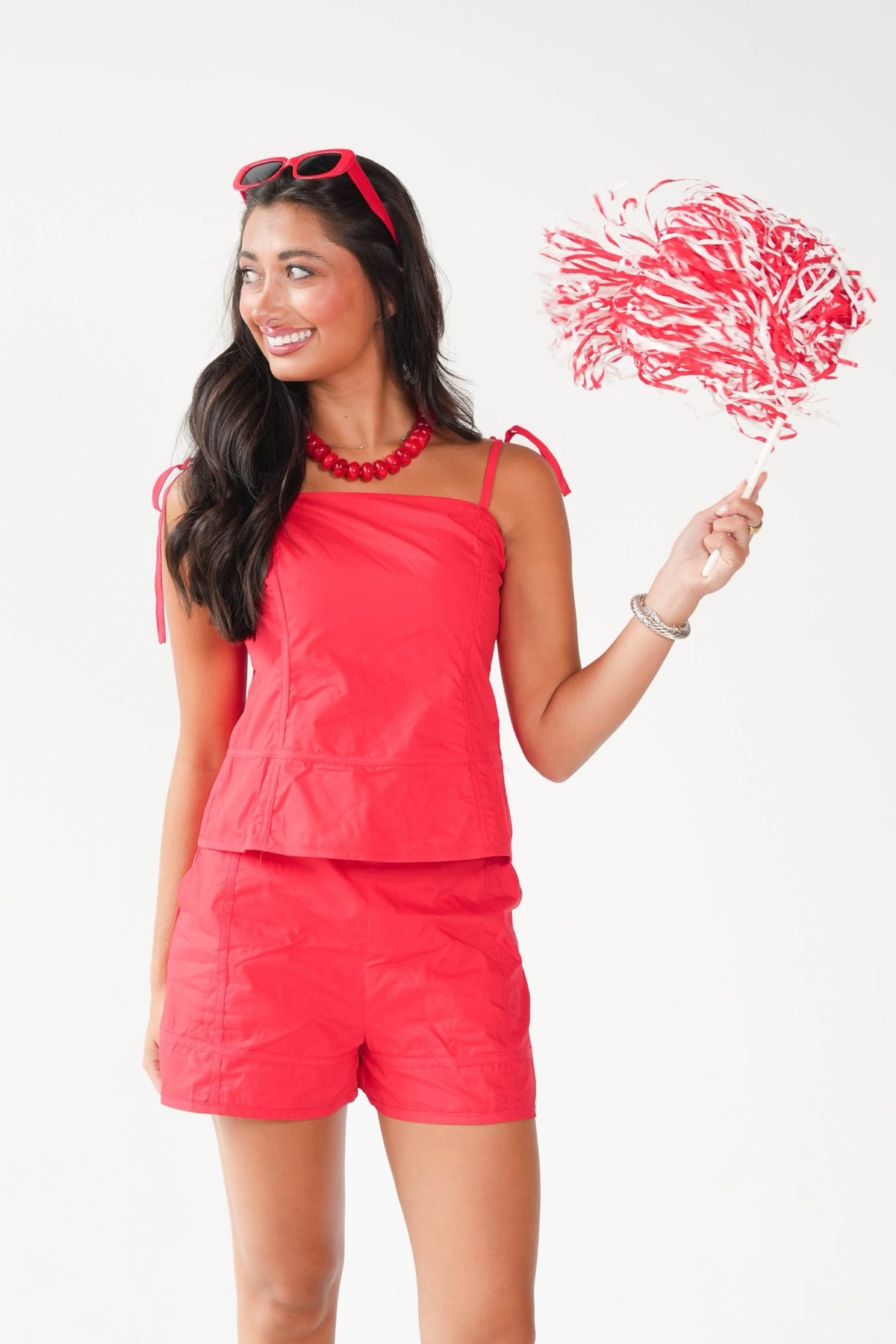 A woman modeling a bright coral-red two-piece outfit consisting of a sleeveless top and matching shorts, holding a red-and-white pom-pom against a plain white background.