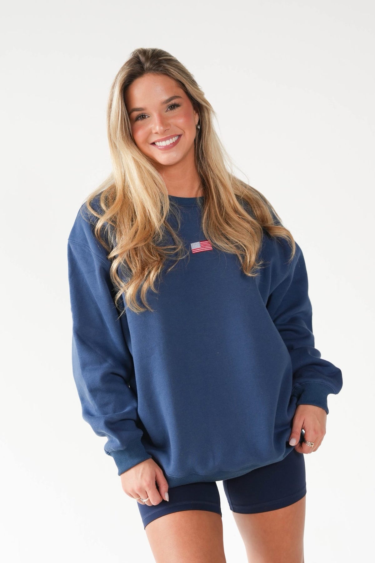 A smiling blonde woman wearing an oversized navy blue crewneck sweatshirt with a small American flag patch on the chest, paired with matching navy bike shorts, against a plain white studio background.