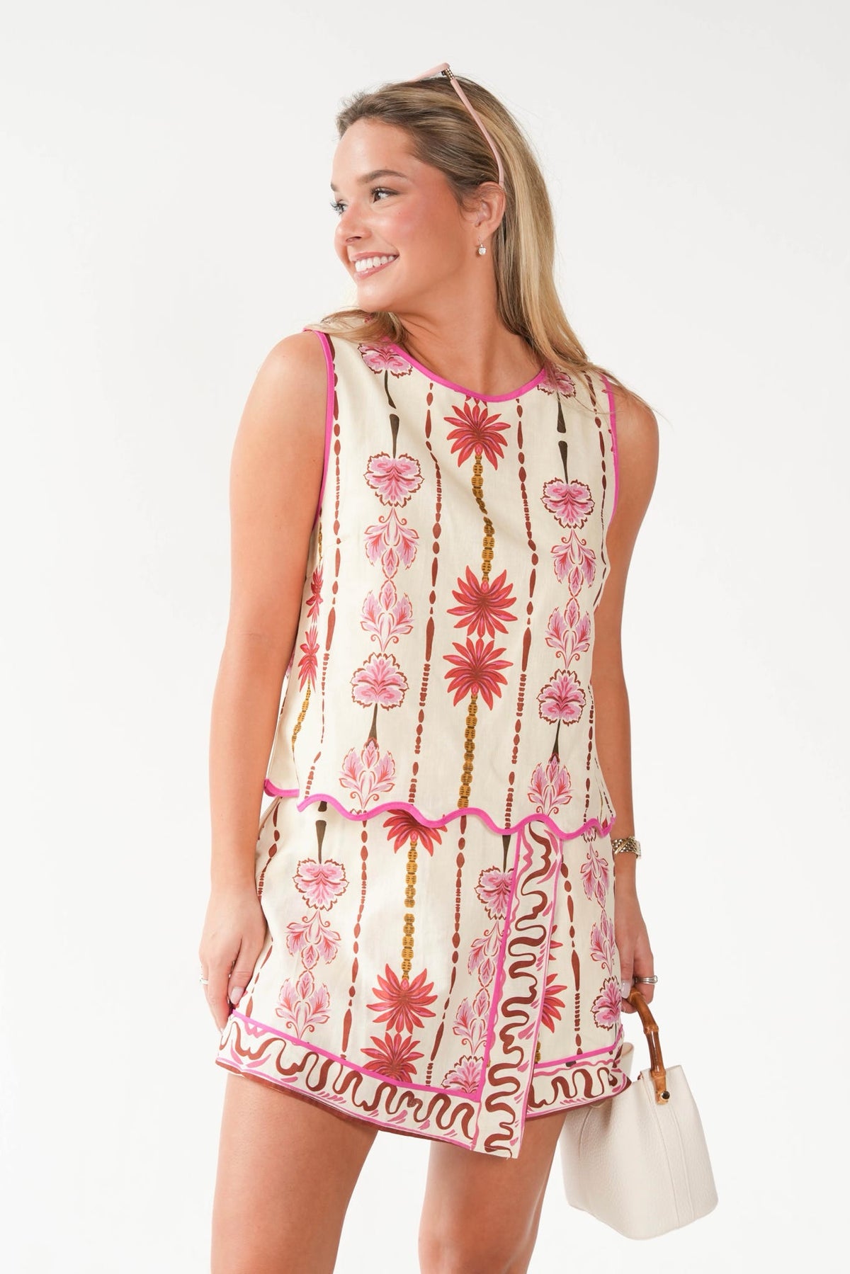 A woman modeling a sleeveless cream-colored mini dress with a vibrant pink and red bohemian-style floral and vertical stripe pattern, holding a small white handbag against a plain white background.