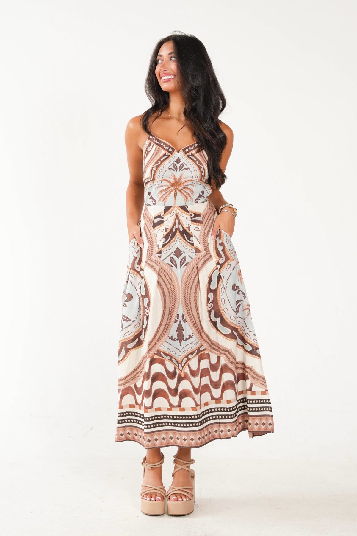 A woman modeling a sleeveless maxi dress with a bohemian-style paisley and floral pattern in beige, brown, and white, paired with beige platform sandals against a plain white studio background.