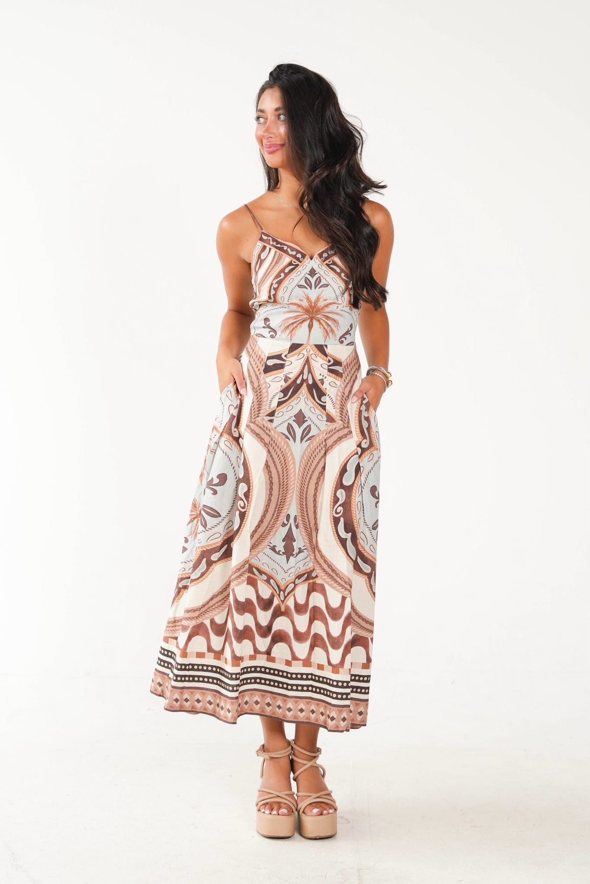 A woman modeling a sleeveless maxi dress with a bohemian-style geometric and floral pattern in beige, brown, and white, paired with tan platform sandals against a plain white studio background.