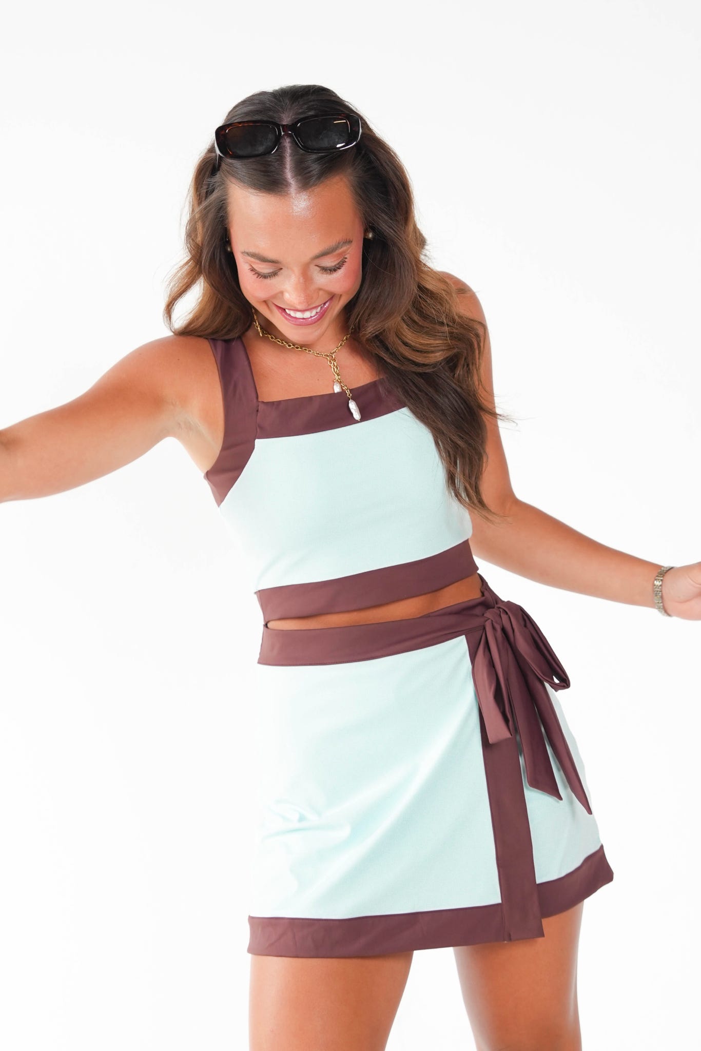 A woman modeling a two-piece athletic set consisting of a light blue crop top and matching mini skirt with dark brown trim, posing against a solid white studio background.