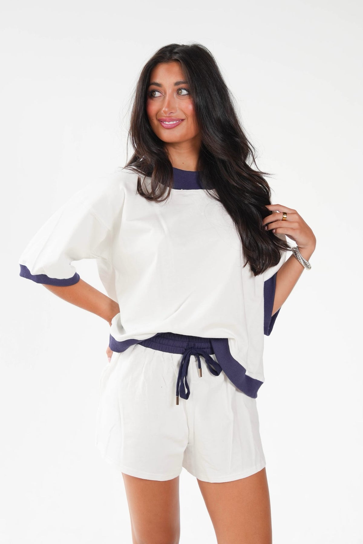 A woman modeling a white two-piece lounge set with navy blue trim, featuring a short-sleeve top with flutter sleeves and matching shorts with a drawstring waist, set against a plain white studio background.