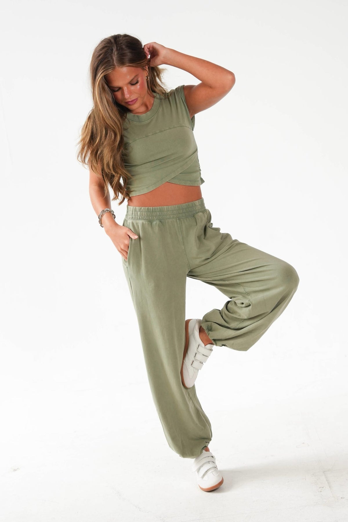 A woman modeling a matching sage green lounge set consisting of a sleeveless crop top and wide-leg sweatpants, paired with white sneakers, against a plain white studio background.
