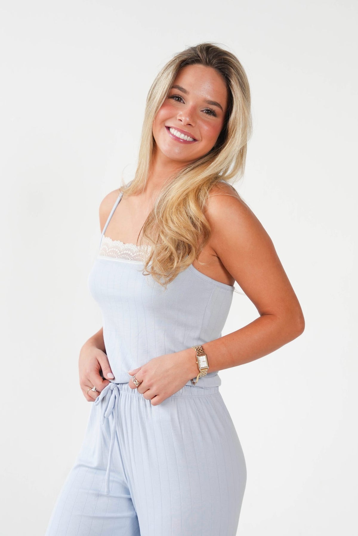 A smiling blonde woman wearing a light blue camisole jumpsuit with a lace-trimmed neckline and a drawstring waist, posed against a clean white studio background.
