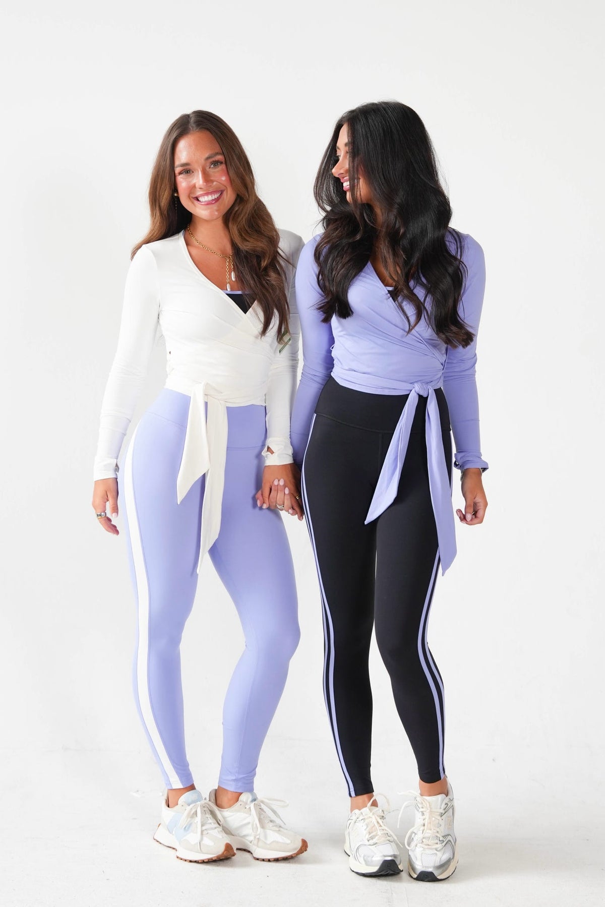 Two women standing side by side in studio, wearing matching wrap-style athletic tops and leggings. The woman on the left wears a white long-sleeve top with light lavender leggings, and the woman on the right wears a light lavender top with black leggings.