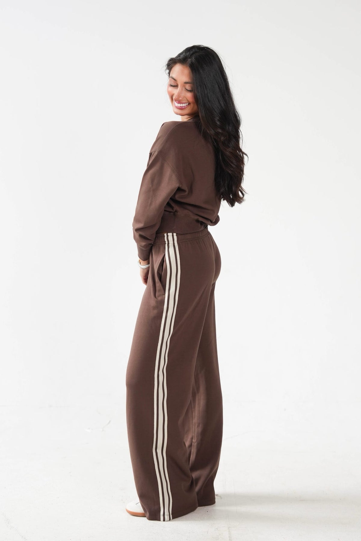A woman wearing a matching brown lounge set with a long-sleeve top and wide-leg pants featuring double cream stripes down the side, photographed against a plain white studio background.